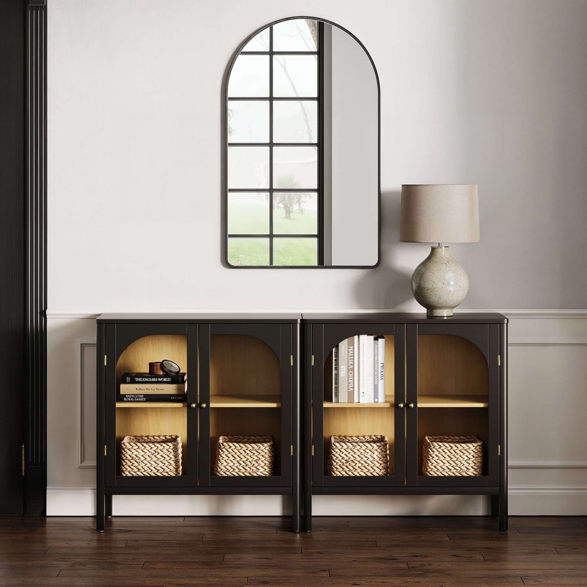 A modern entryway features two Nathan James Curio Cabinet TV Stand Credenzas with glass doors, black finish, and wicker baskets inside, topped with a beige lamp and an arched mirror above, set against a white wainscoted wall.