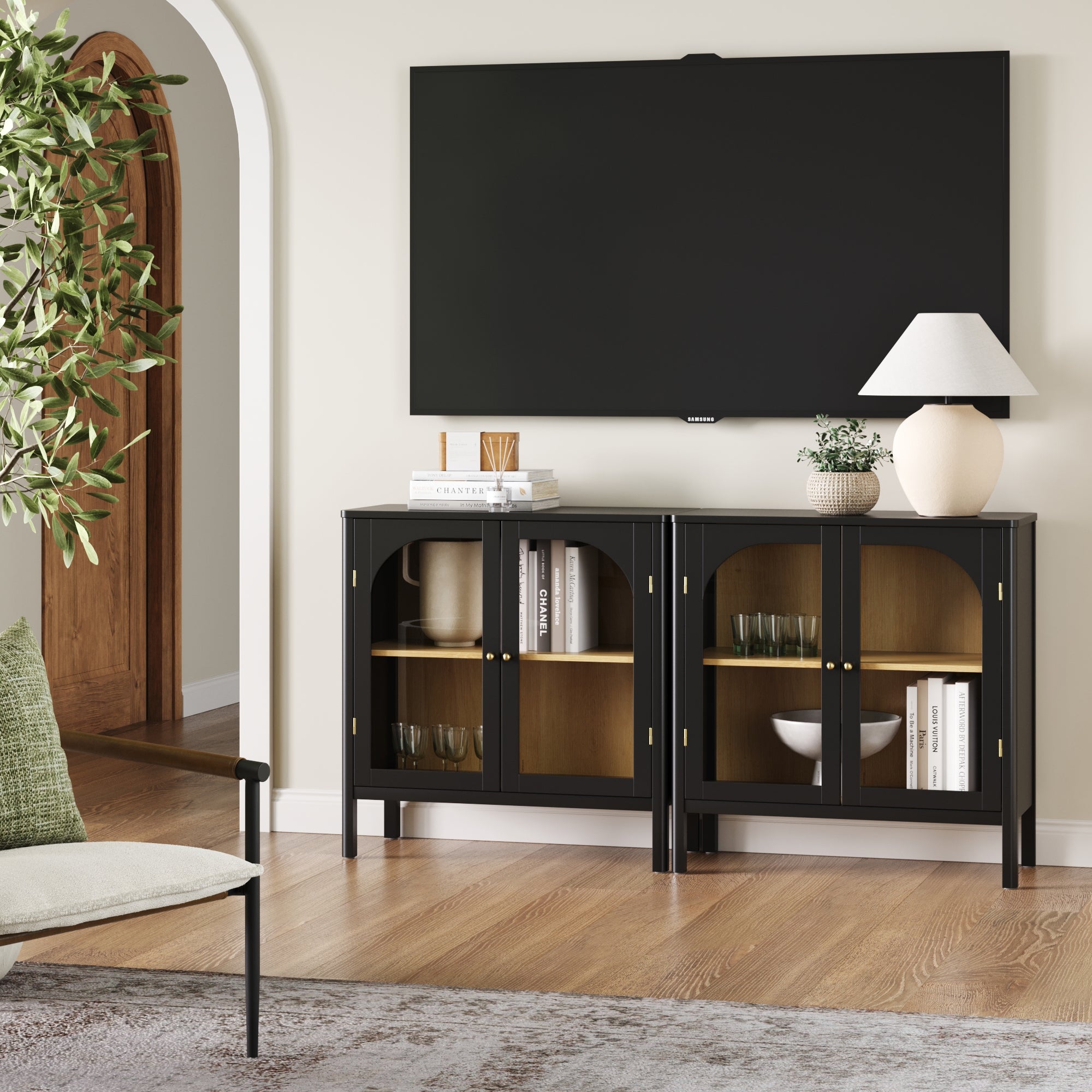 A modern living room features a Nathan James Curio Cabinet TV Stand Credenza with Glass Door Black (Set of 2) beneath a wall-mounted TV. Decorative items, a white lamp, vase, and books adorn the top. A chair and green plant are visible in the foreground.