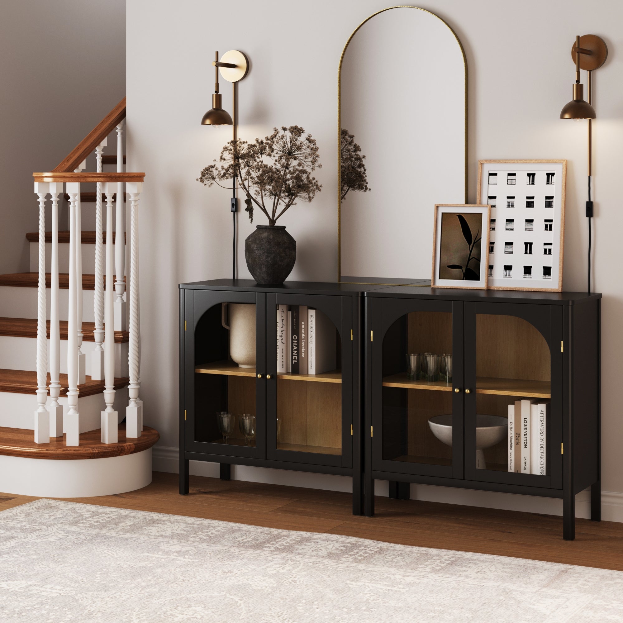 A modern entryway features two Nathan James Curio Cabinet TV Stand Credenzas with Glass Doors in black, storing books and decor, beside a large arched mirror, wall sconces, a vase of dried branches, and a white staircase with wooden steps.