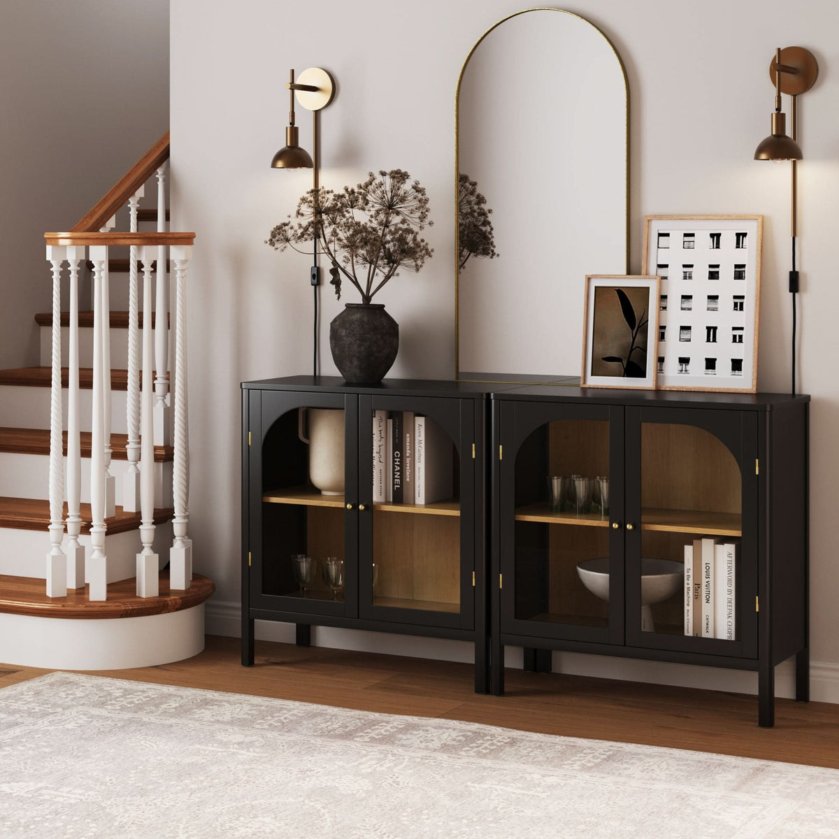 A set of two Nathan James Curio Cabinet Credenza with Glass Door Black, featuring solid wood, arched glass doors, and brass hardware, placed side-by-side beneath a gold-framed mirror and two wall sconces.