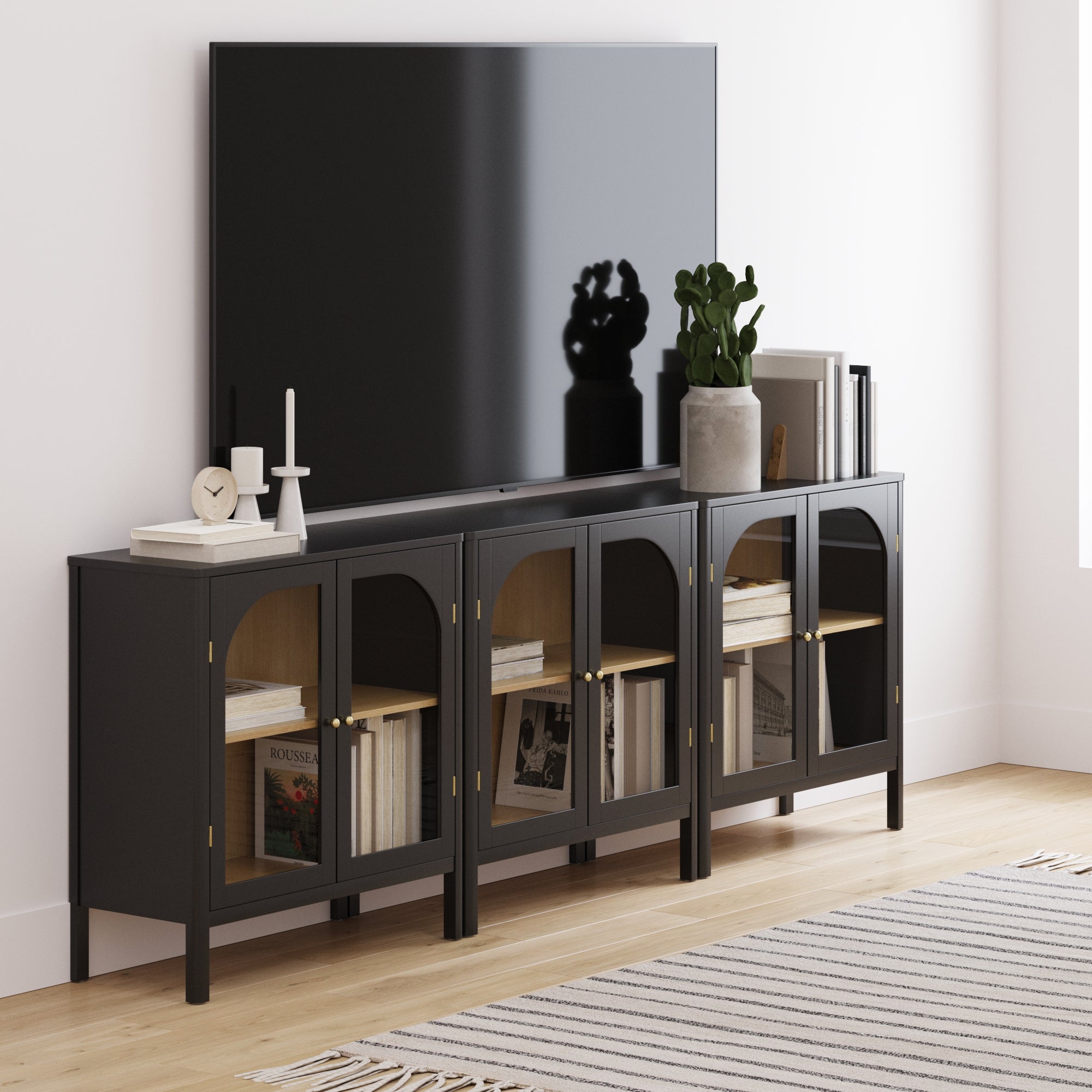 A modern living room features a large TV above the Nathan James Curio Cabinet TV Stand Credenza with Glass Door Black (Set of 3), styled with books, plants, candles, and decor on a light wood floor with a striped rug.