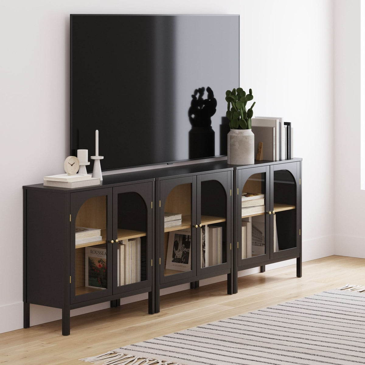 Three Nathan James Curio Cabinet Credenza with Glass Door Black storage cabinets with arched glass doors and brass finish hardware arranged side-by-side to support a wall-mounted television.