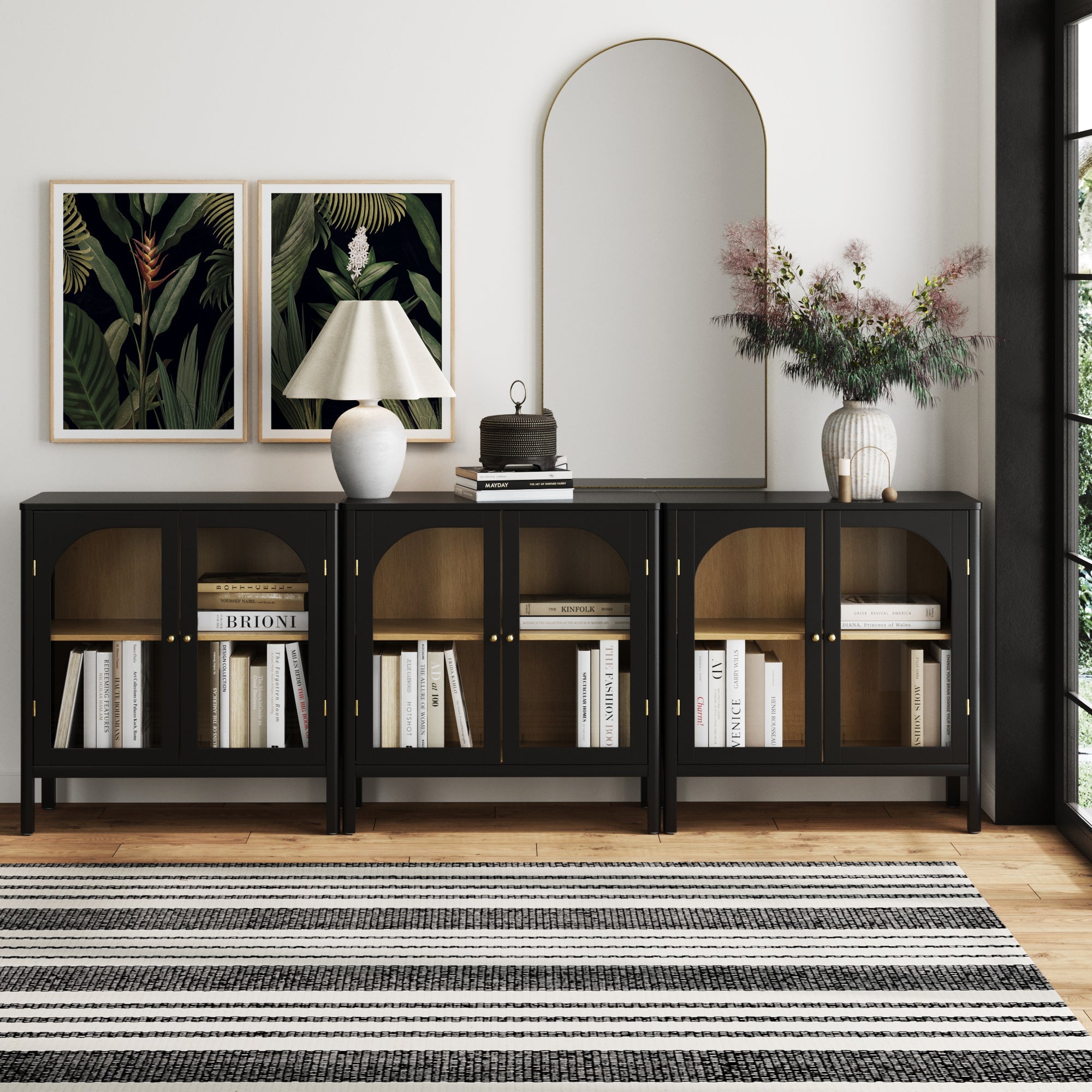 A modern living room showcases the Nathan James Curio Cabinet TV Stand Credenza with Glass Door Black (Set of 3), styled with books, a lamp, and vases. Two botanical art prints and an arched mirror hang above, while a striped rug covers the wooden floor.