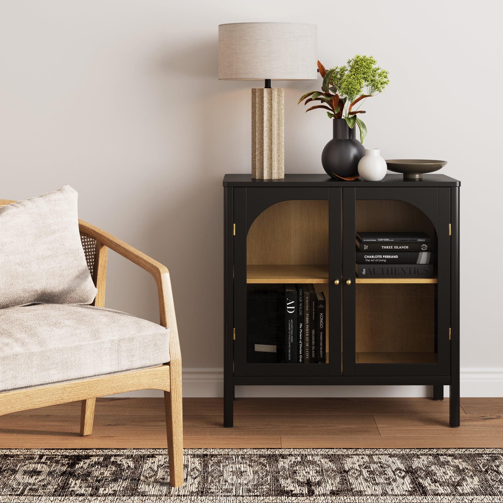 A modern living room features a wooden armchair and the Nathan James Wood Curio Cabinet with Glass Doors in Black, accented by brass hardware. Books, decor, a lamp, vase, bowl, and greenery complement the setup against a white wall and patterned rug.