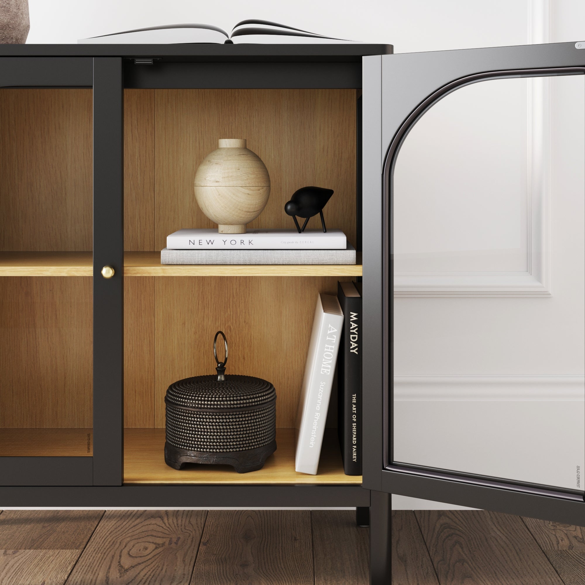 A black cabinet with wooden shelves holds a round beige vase, a small black bird figurine, books, and a dark textured container, with an open book resting on top and the cabinet door open.