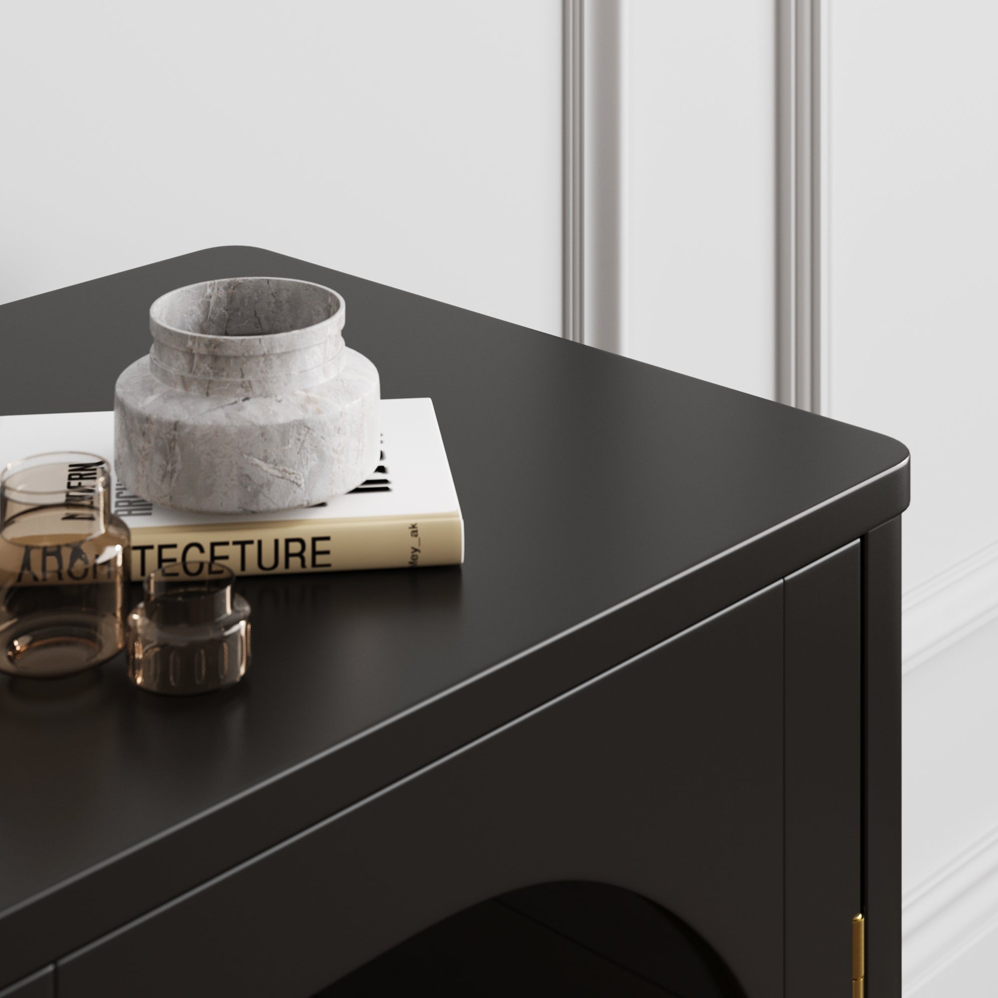 A close-up of a black cabinet with a marble vase, two glass cups, and a book titled ARCHITECTURE placed on top, set against a white wall with decorative molding.