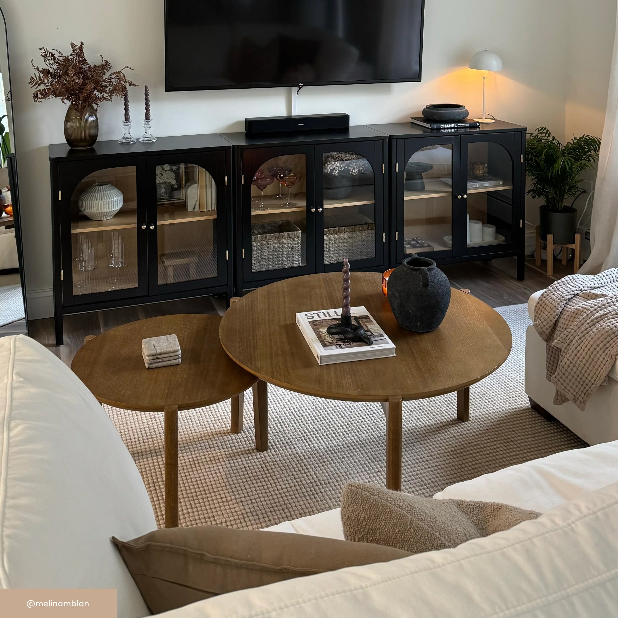A cozy living room with a white sofa, two round wooden coffee tables holding decor items, a black cabinet with glass doors, a TV mounted above, and neutral-toned accents throughout the room.
