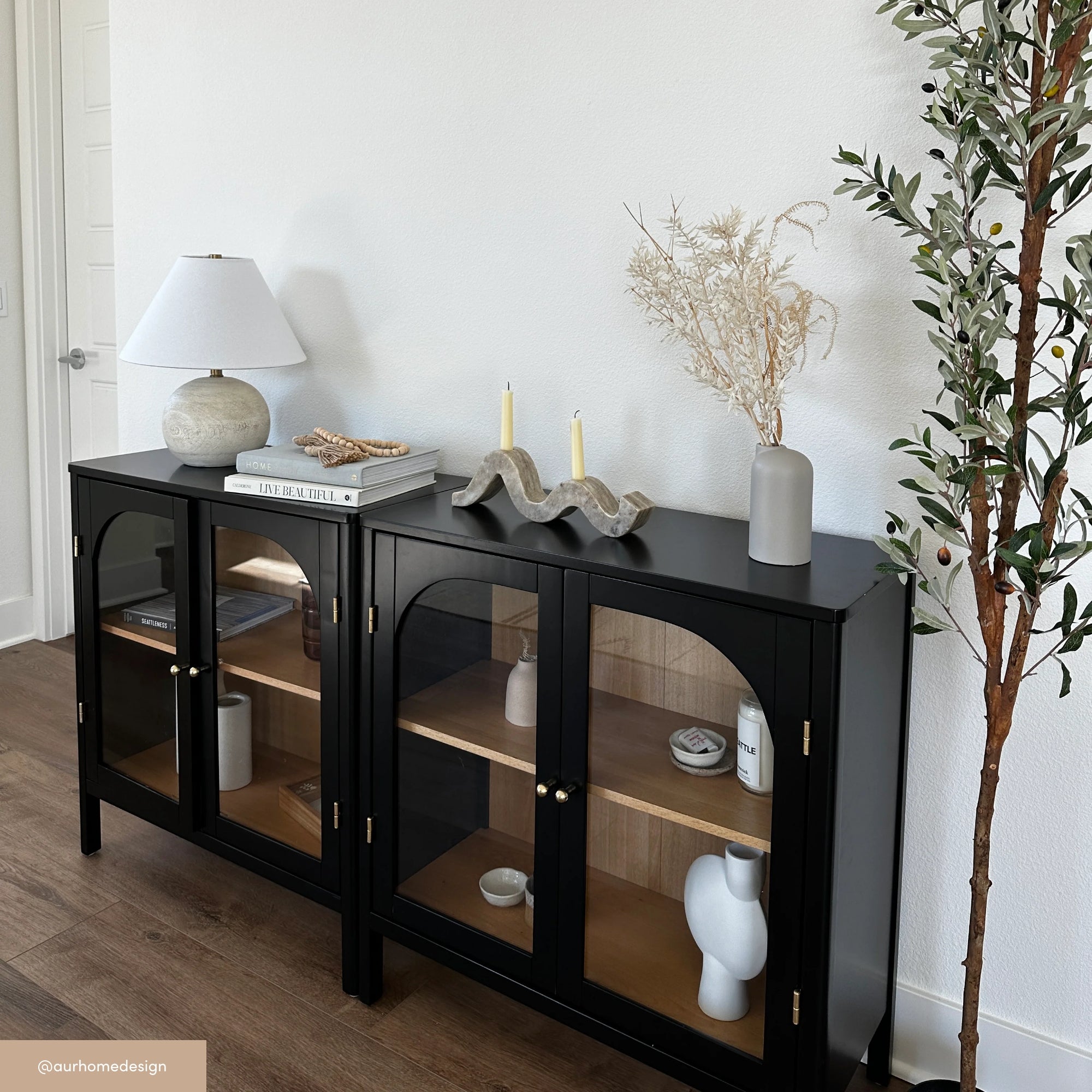 Two black display cabinets with glass doors sit against a white wall. On top are decorative items: a lamp, books, candles, a ceramic vase, and dried foliage. A potted faux olive tree stands next to the cabinets.