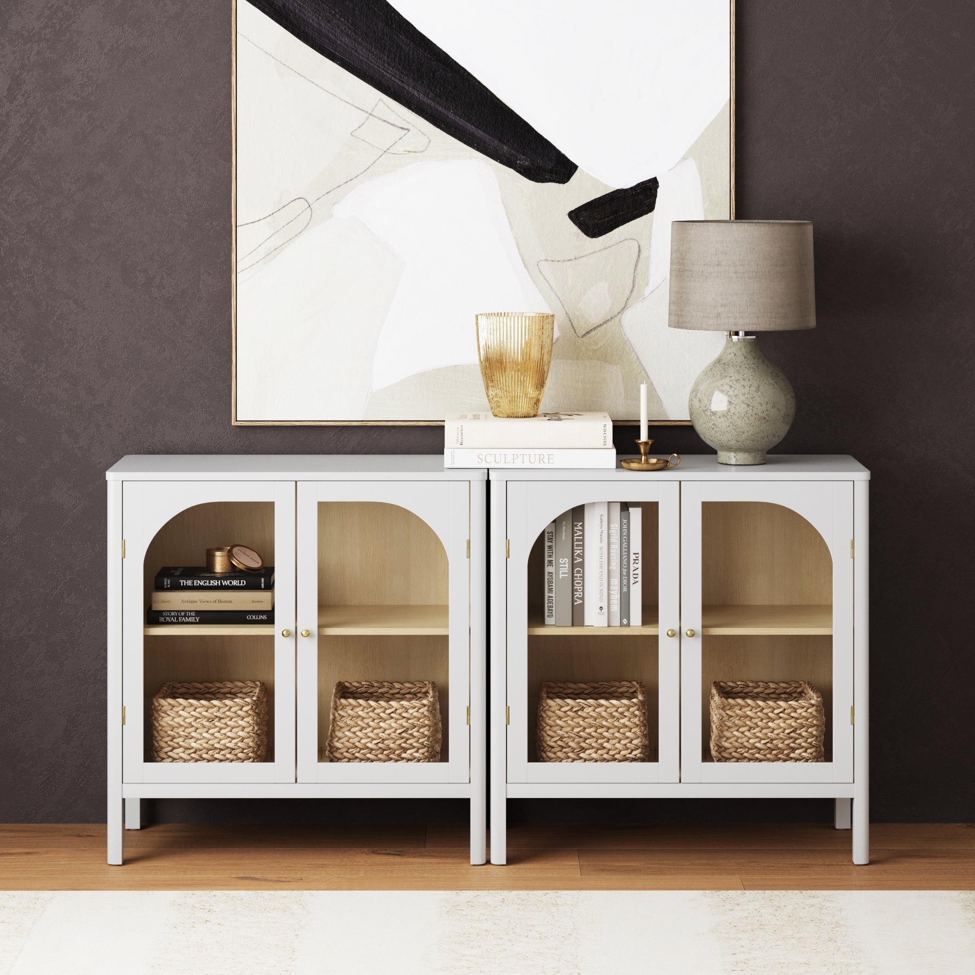 The Nathan James Curio Cabinet TV Stand Credenza with Glass Door in White (Set of 2) displays woven baskets and books behind curved glass doors with brass hardware. A lamp, glass, book, and candle sit on top; an abstract painting hangs above on a dark wall.