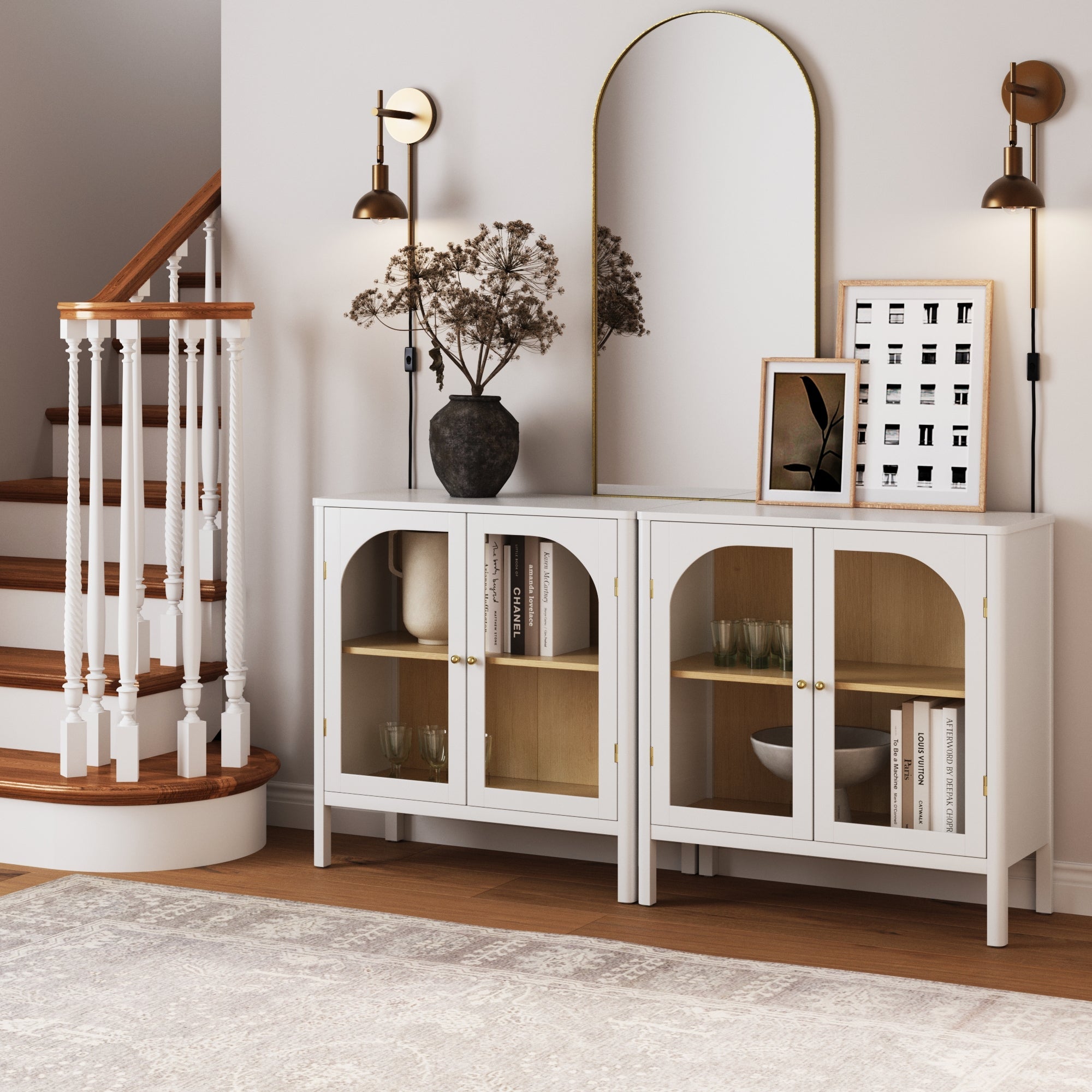 A stylish entryway showcases two Nathan James Curio Cabinet TV Stand Credenzas with glass doors, books, decor pieces, a dark vase with dried branches, framed art, a gold-framed mirror, sconces, and a wooden staircase.