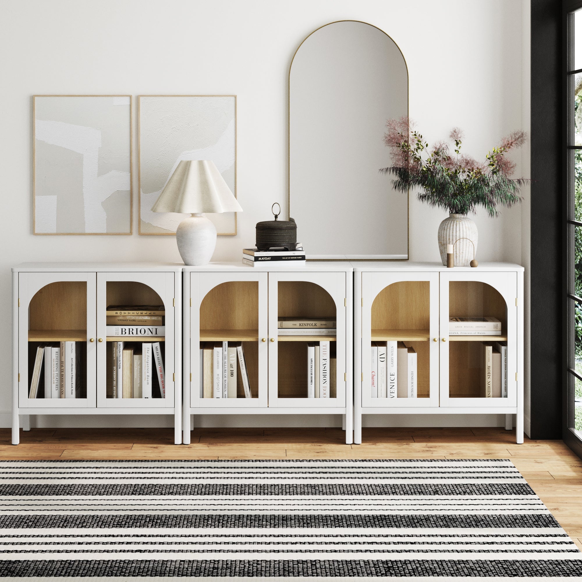 A modern living room features the Nathan James Curio Cabinet TV Stand Credenza with Glass Door in white (Set of 3), styled with books, decor, a table lamp, large mirror, abstract wall art, and a vase on a black and white striped rug.