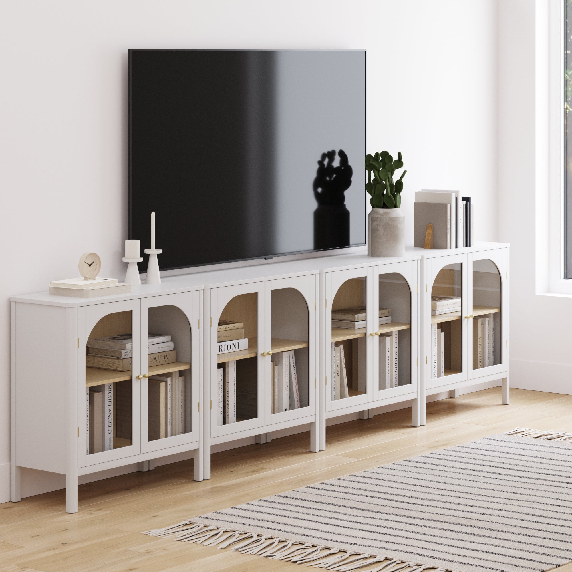 A modern living room showcases a wall-mounted flat-screen TV above the Nathan James Large Glass Door Curio Cabinet TV Console Credenza White (Set of 4), styled with books, candles, a plant, brass hardware, and a striped rug on light wood floors.
