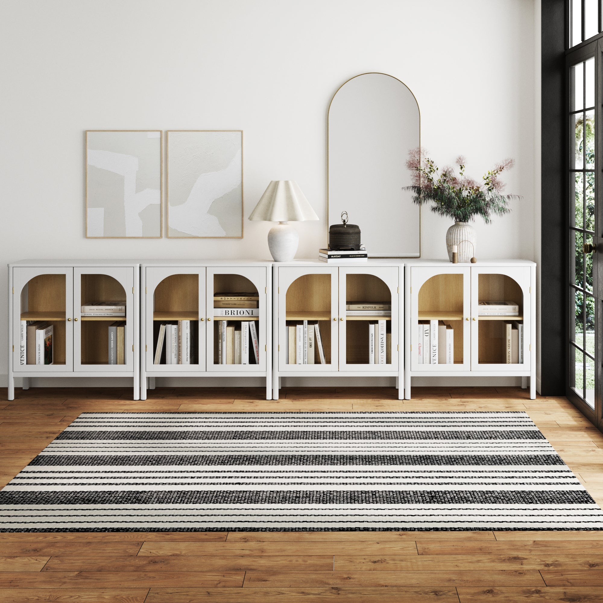 A modern living room showcases the Nathan James Large Glass Door Curio Cabinet TV Console Credenza in white (set of 4), accented with books, decor, abstract art, a large arched mirror, lamp, flowers in a vase, and a black-and-white striped rug on wood floors.