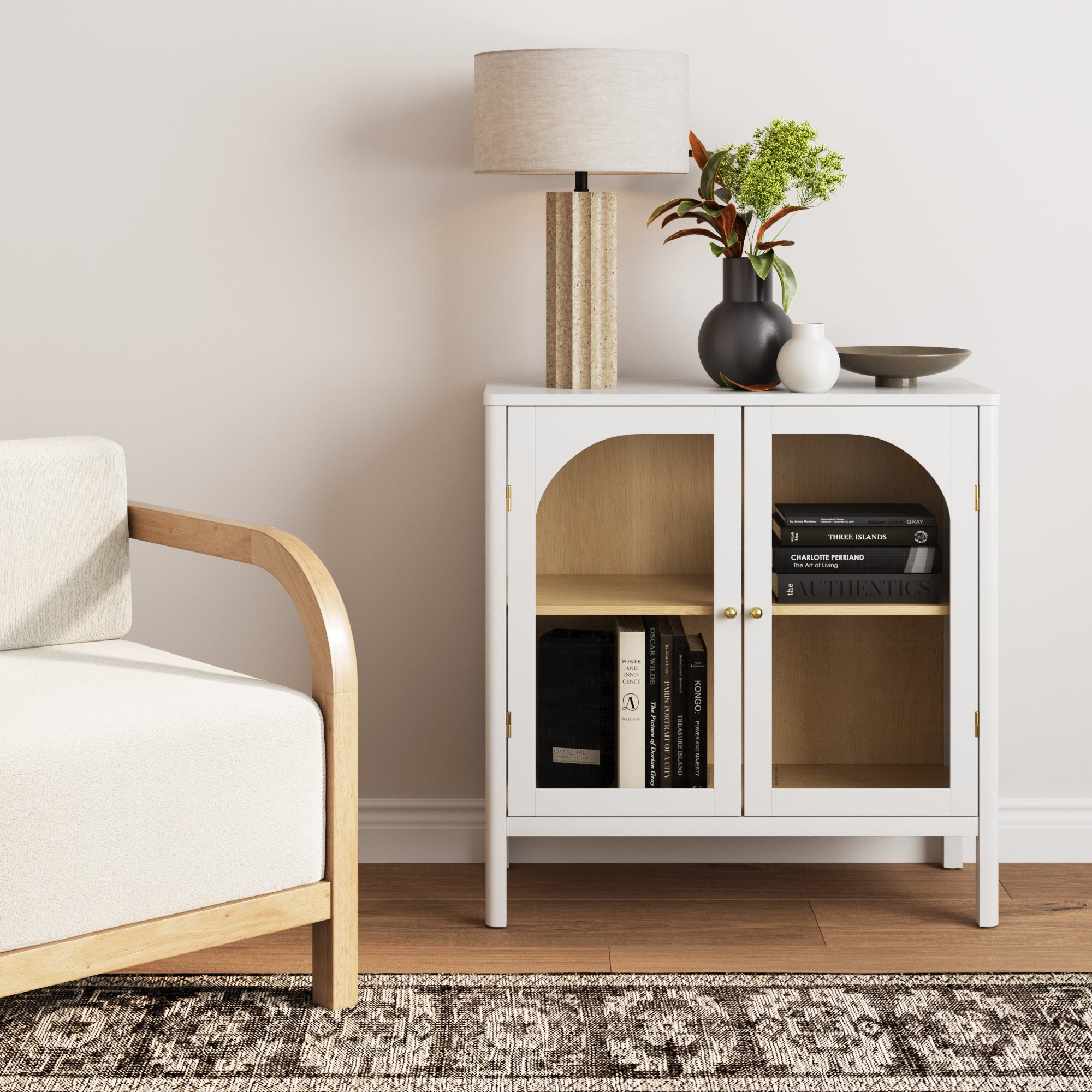 A modern living room corner featuring the Nathan James Wood Curio Cabinet with Glass Doors in white, a beige armchair, patterned rug, table lamp, and decorative vases with greenery on top of the cabinet.