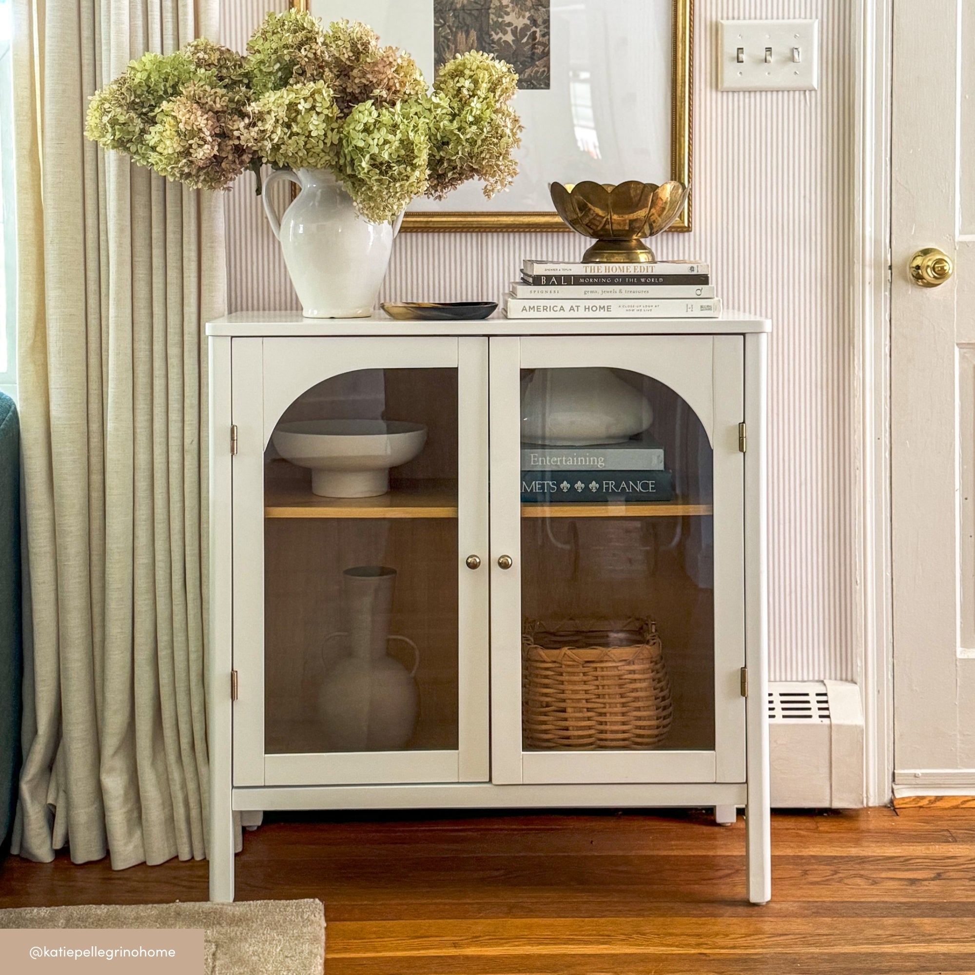 A white cabinet with glass doors holds books and a wicker basket. On top, theres a large vase with dried flowers, stacked books, and a gold bowl. The cabinet sits on a wooden floor next to a door and light-colored curtain.