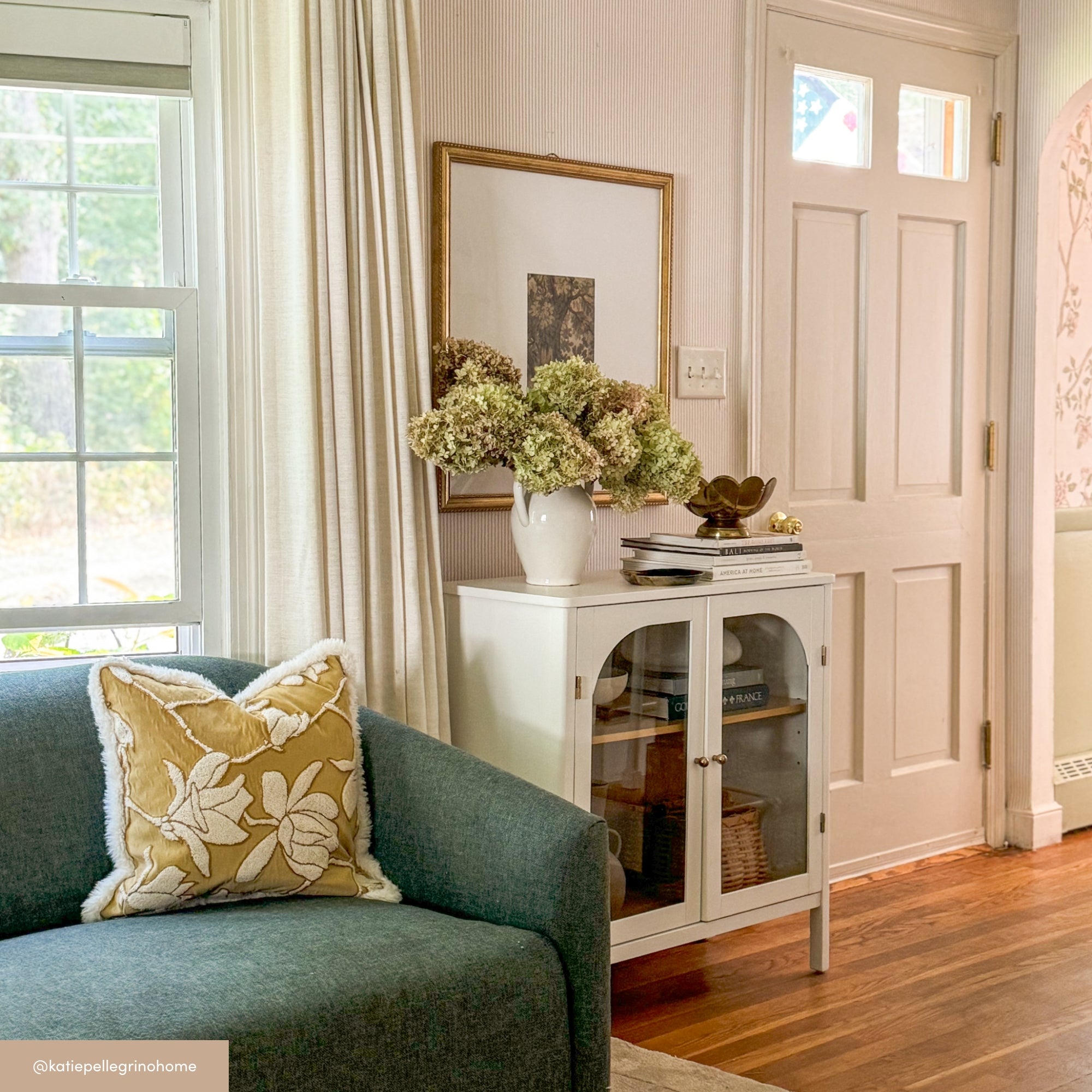 A cozy living room corner with a green sofa, a yellow floral pillow, white curtains, a window, a white cabinet with books and decor, a vase of dried flowers, framed art, and a white front door.