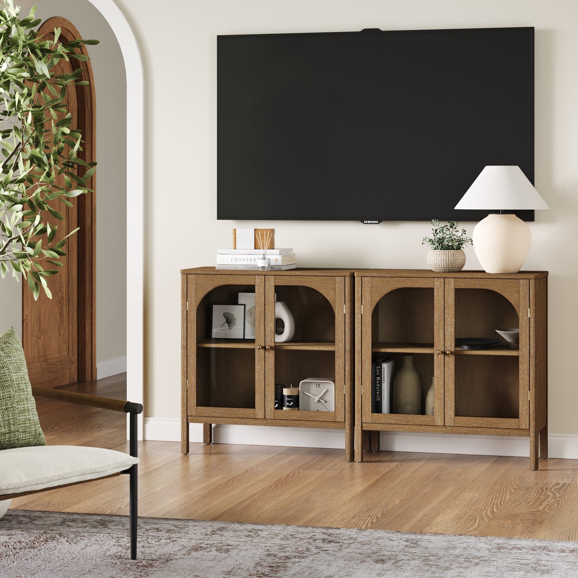 A modern living room showcases a wall-mounted TV above two Nathan James Curio Cabinet TV Stand Credenzas with glass doors and brass hardware, displaying books and decor. A leafy plant is beside a cushioned chair in the foreground.