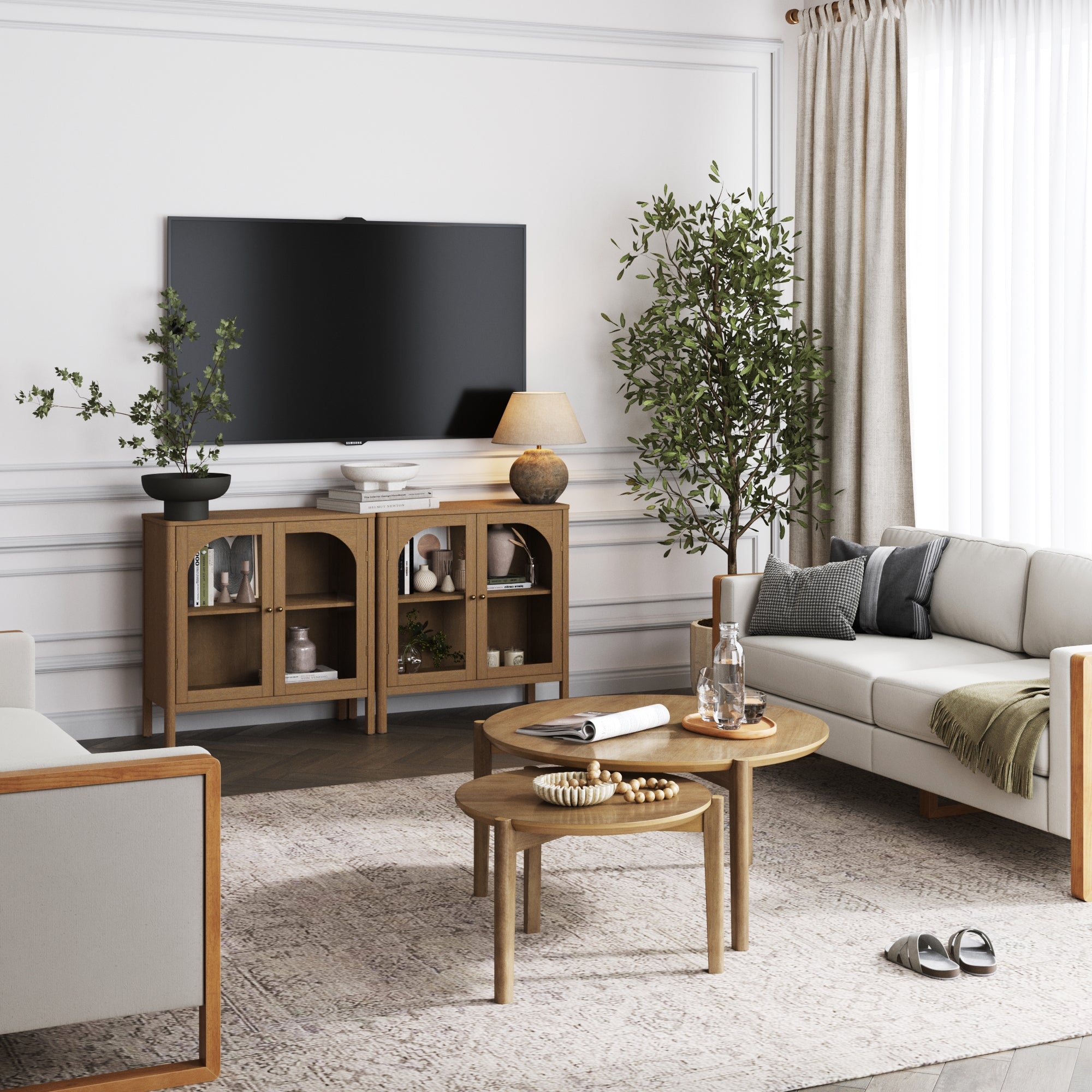 A modern living room with a wall-mounted TV, wooden cabinet with decor items, two round wooden coffee tables, neutral sofas, potted plants, and large windows with beige curtains. Natural light brightens the space.