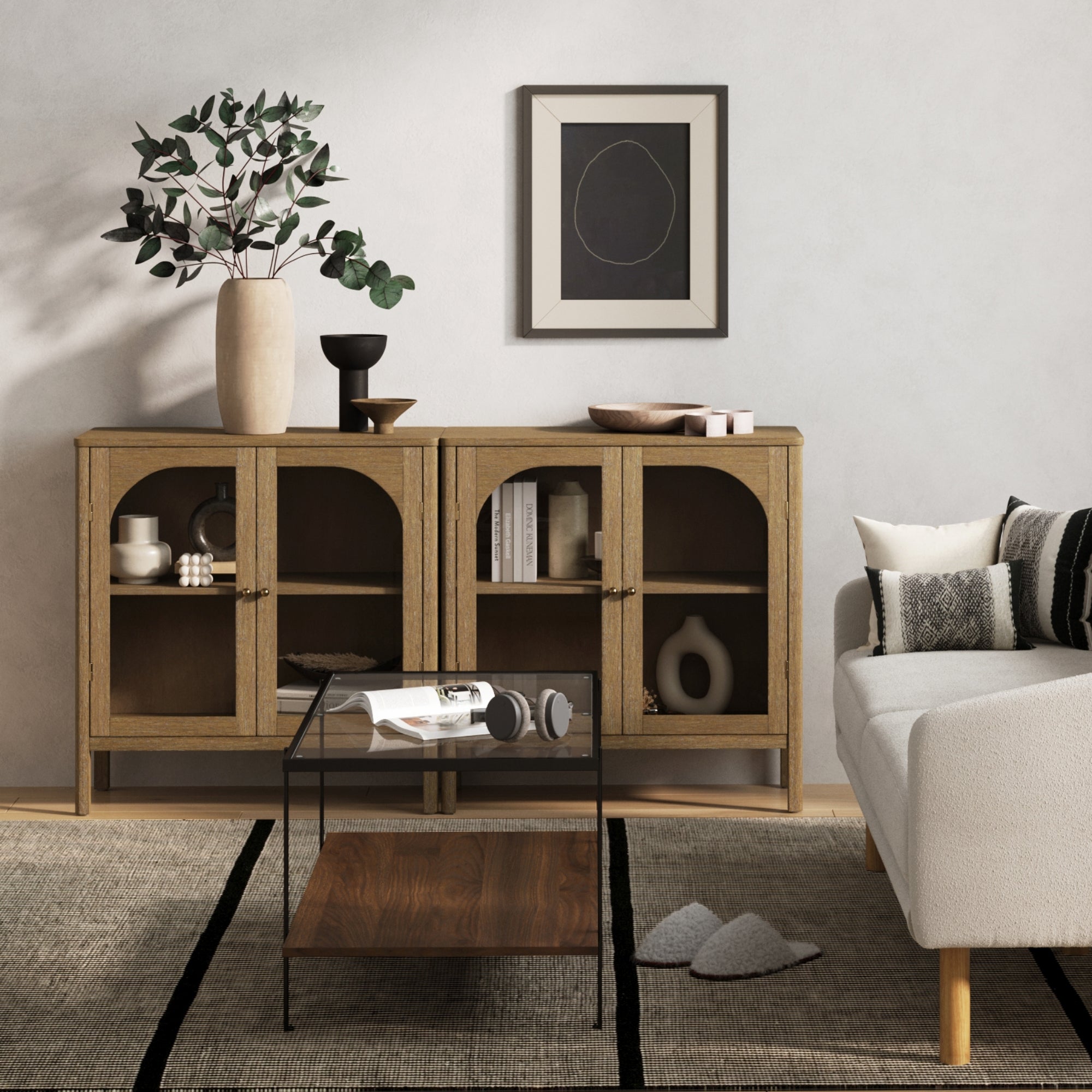 A modern living room with two Nathan James Curio Cabinet TV Stand Credenzas in brown with glass doors, brass hardware, decorative vases, abstract art, a white sofa with patterned cushions, a small glass coffee table, and a neutral rug.