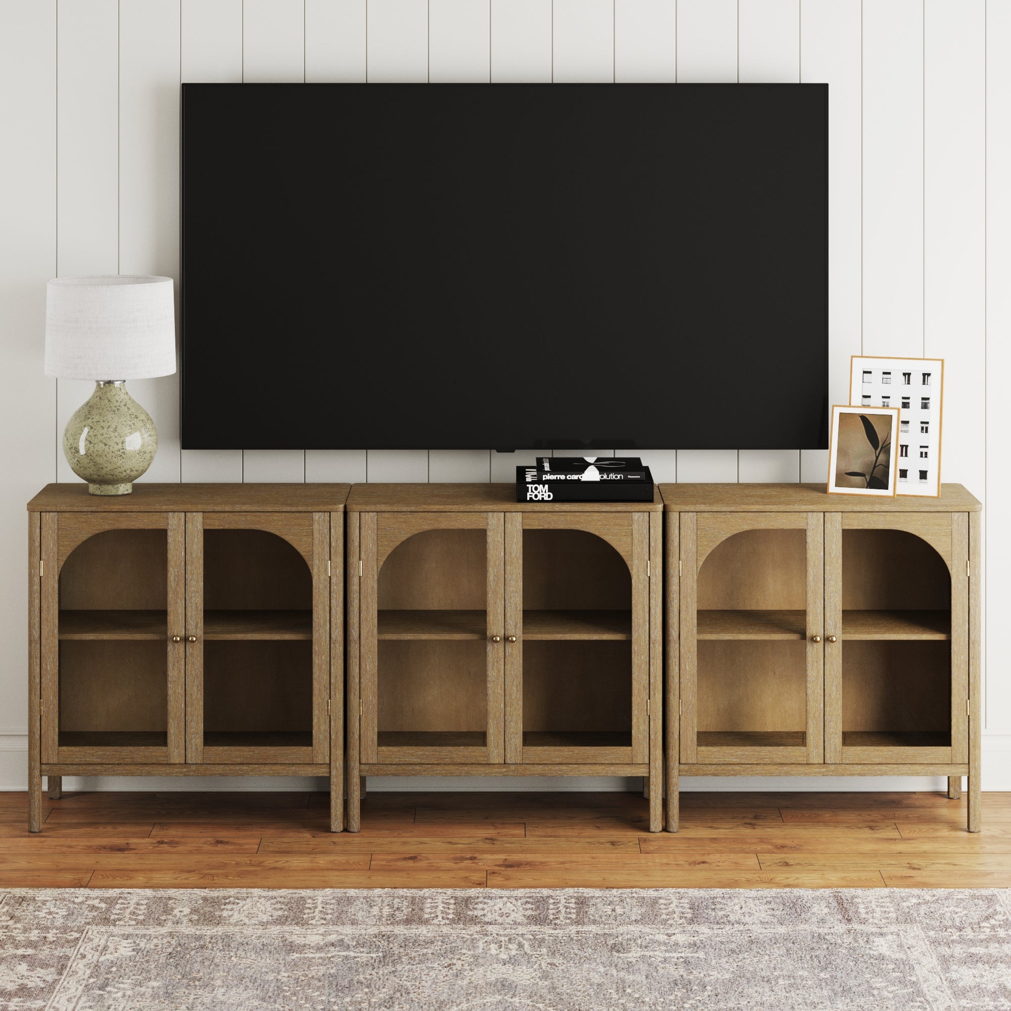A modern living room features the Nathan James Curio Cabinet TV Stand Credenza with Glass Door Brown (Set of 3) below a large black wall-mounted TV, green lamp, stereo, and framed art on wood floors with a patterned rug.