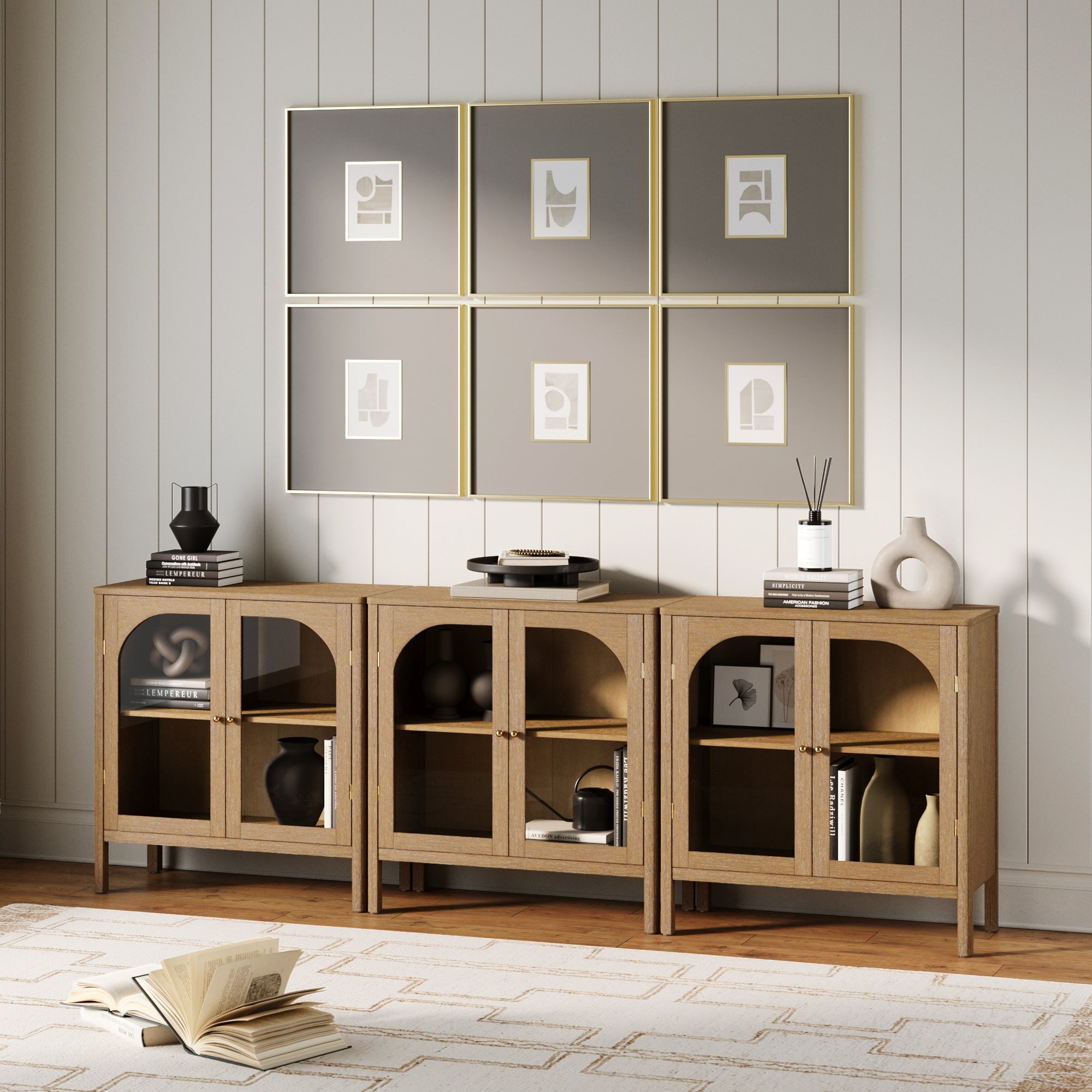 A modern living room showcases three Nathan James Curio Cabinet TV Stand Credenzas with glass doors in brown, brass hardware, minimalist accents, and six abstract framed artworks above on a white shiplap wall. An open book and rug sit on the floor.