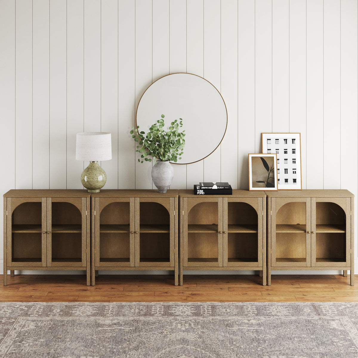 A set of four Nathan James Large Glass Door Curio Cabinet Credenza Brown units with solid wood legs and arched glass doors arranged against a white wall, featuring decor, brass hardware, and a circular wall mirror above.