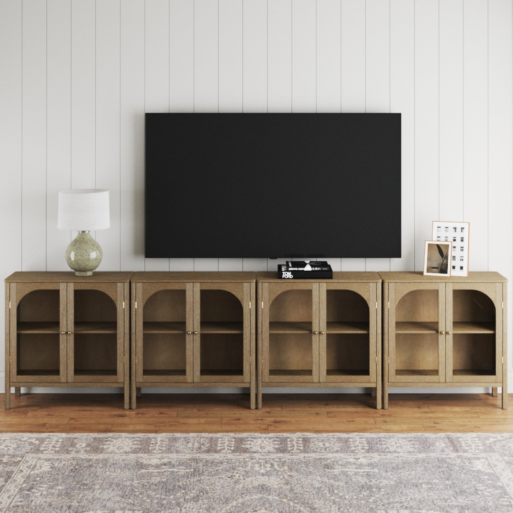 A large TV is mounted on a white shiplap wall above the Nathan James Large Glass Door Curio Cabinet TV Console Credenza in brown, which displays a lamp, books, framed photo, and decor. A patterned rug is spread across the floor.