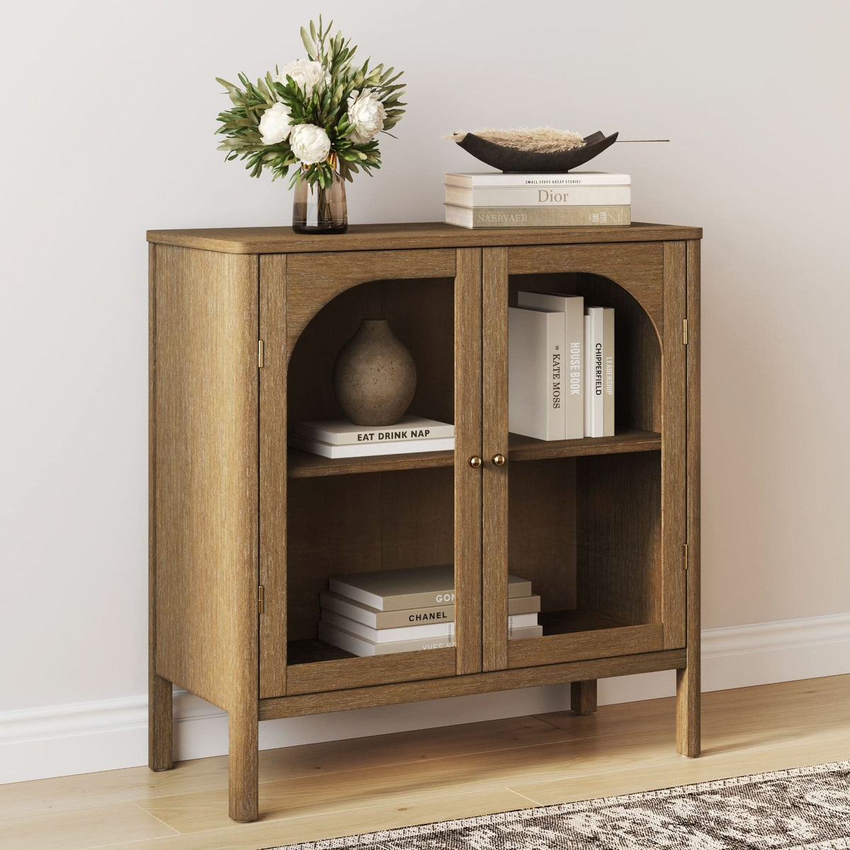 The Nathan James Wood Curio Cabinet with Glass Doors in brown displays books and a vase inside. On top, a bowl, stacked books, and a vase with white flowers sit. The solid wood cabinet stands against a light wall on hardwood flooring.