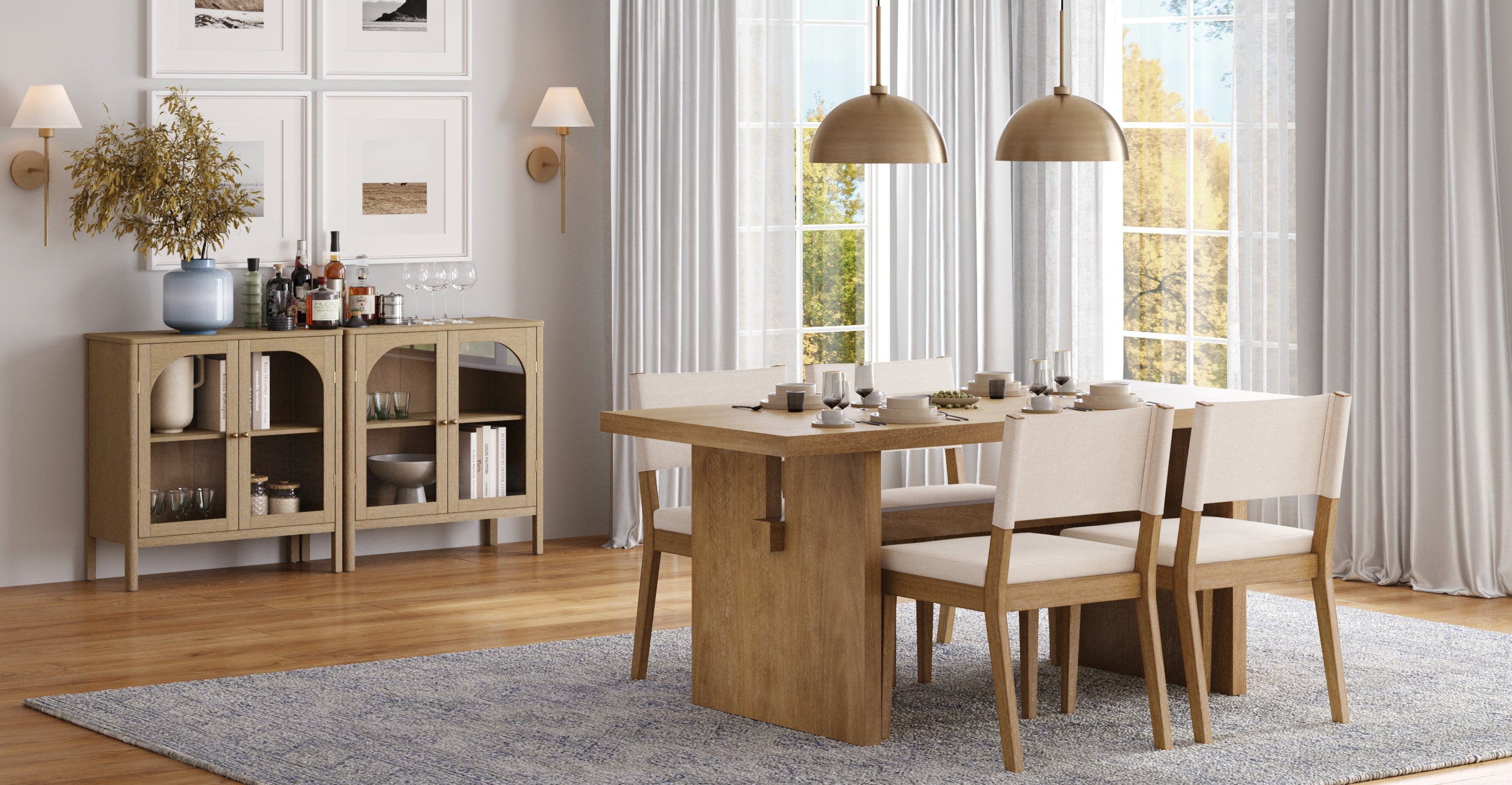 A modern dining room with a wooden table, four cushioned chairs, a sideboard with decor and drinks, pendant lights, framed art, and large windows with sheer curtains letting in natural light.