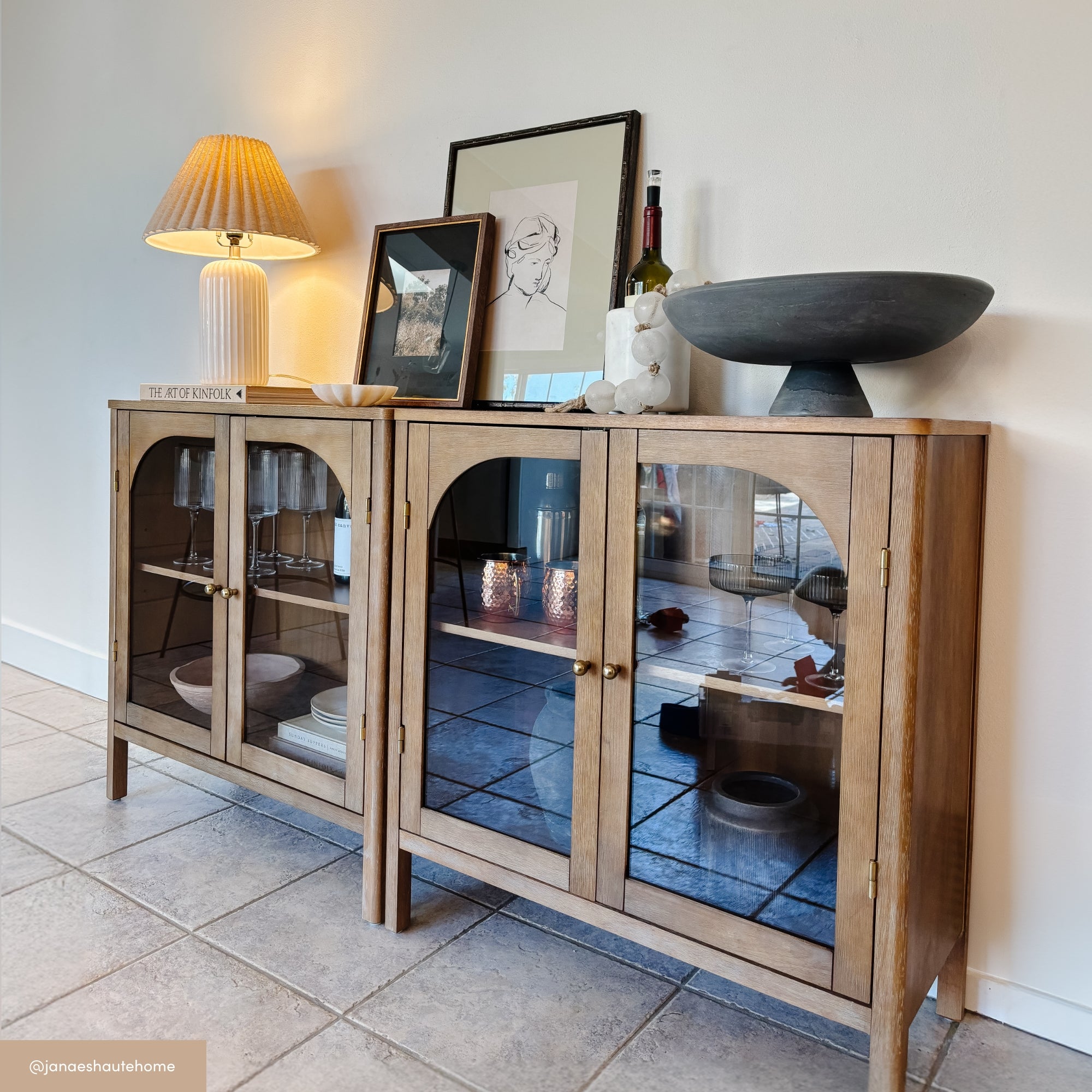 A wooden glass-front sideboard is decorated with a table lamp, framed artwork, a bowl, wine bottle, and candles. Reflections of decor appear in the glass doors. The scene sits on a tiled floor against a white wall.