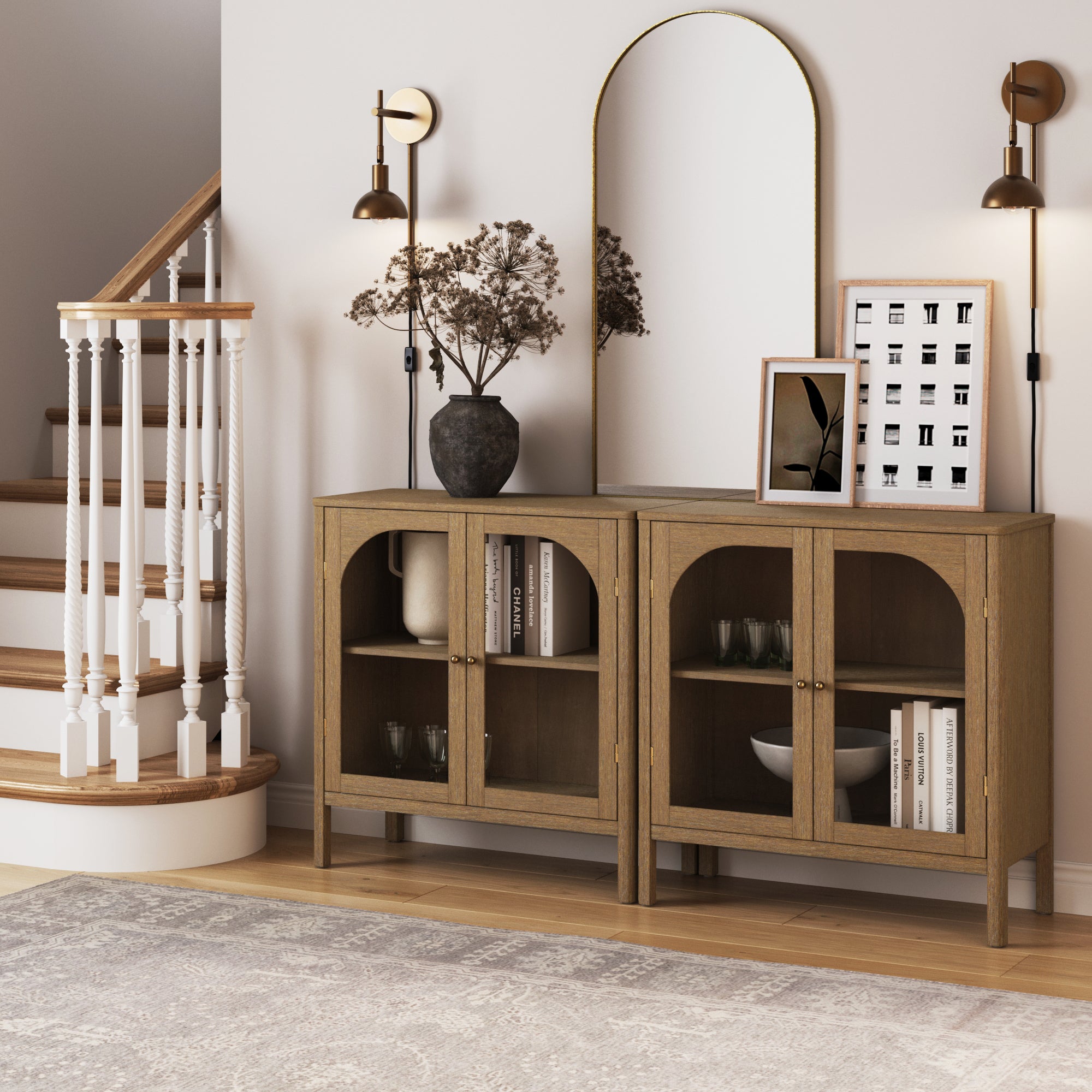 The Nathan James Curio Cabinet TV Stand Credenza with Glass Door in brown (set of 2) stands against a white wall, topped with vases, books, framed art, and a round mirror. Two wall sconces and a staircase are beside a patterned rug.