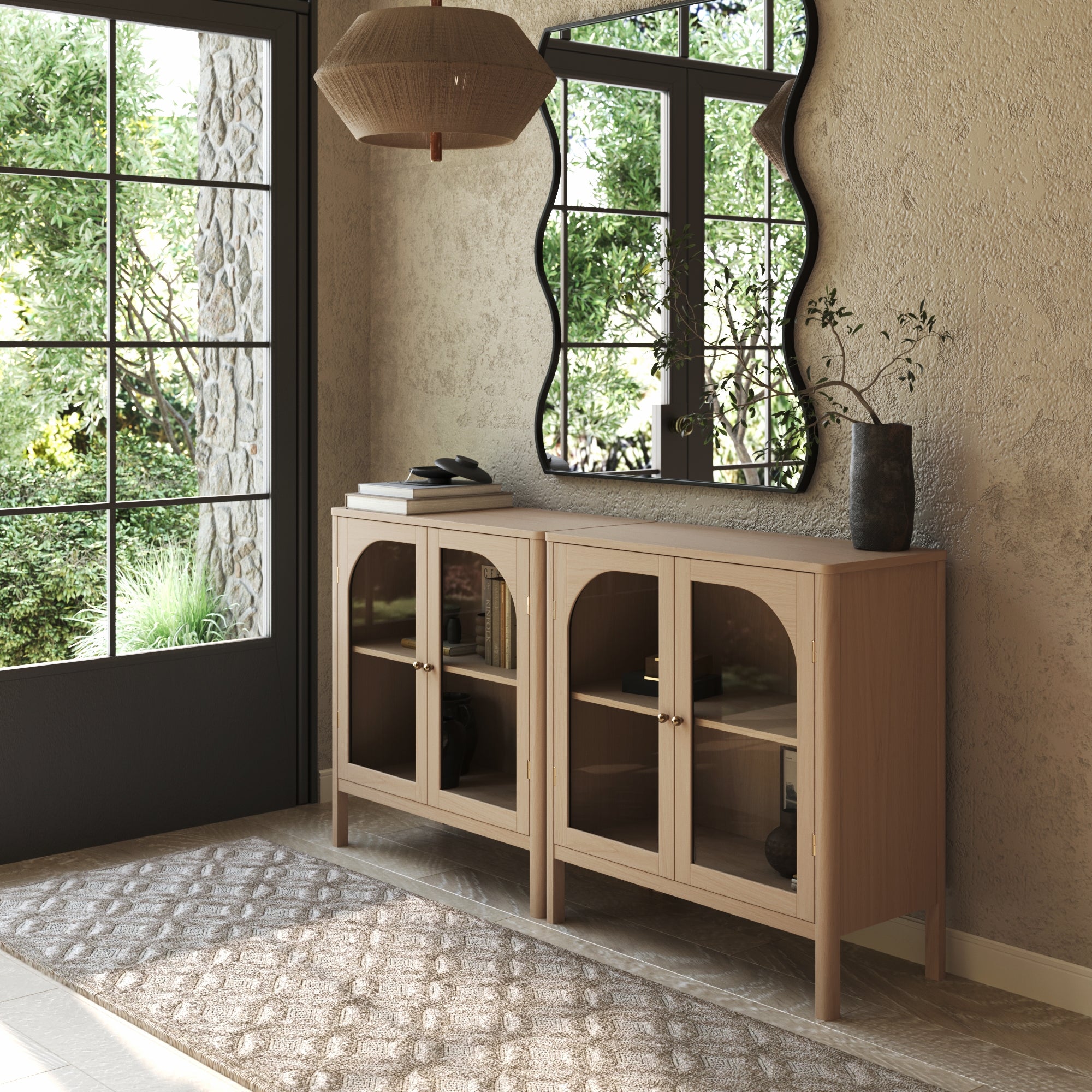 A stylish entryway is anchored by a set of two Nathan James Curio Cabinet TV Stand Credenzas in Raw Oak with glass doors, paired with a wavy mirror, vase of branches, patterned neutral rug, and large windows that welcome natural light and outdoor greenery.