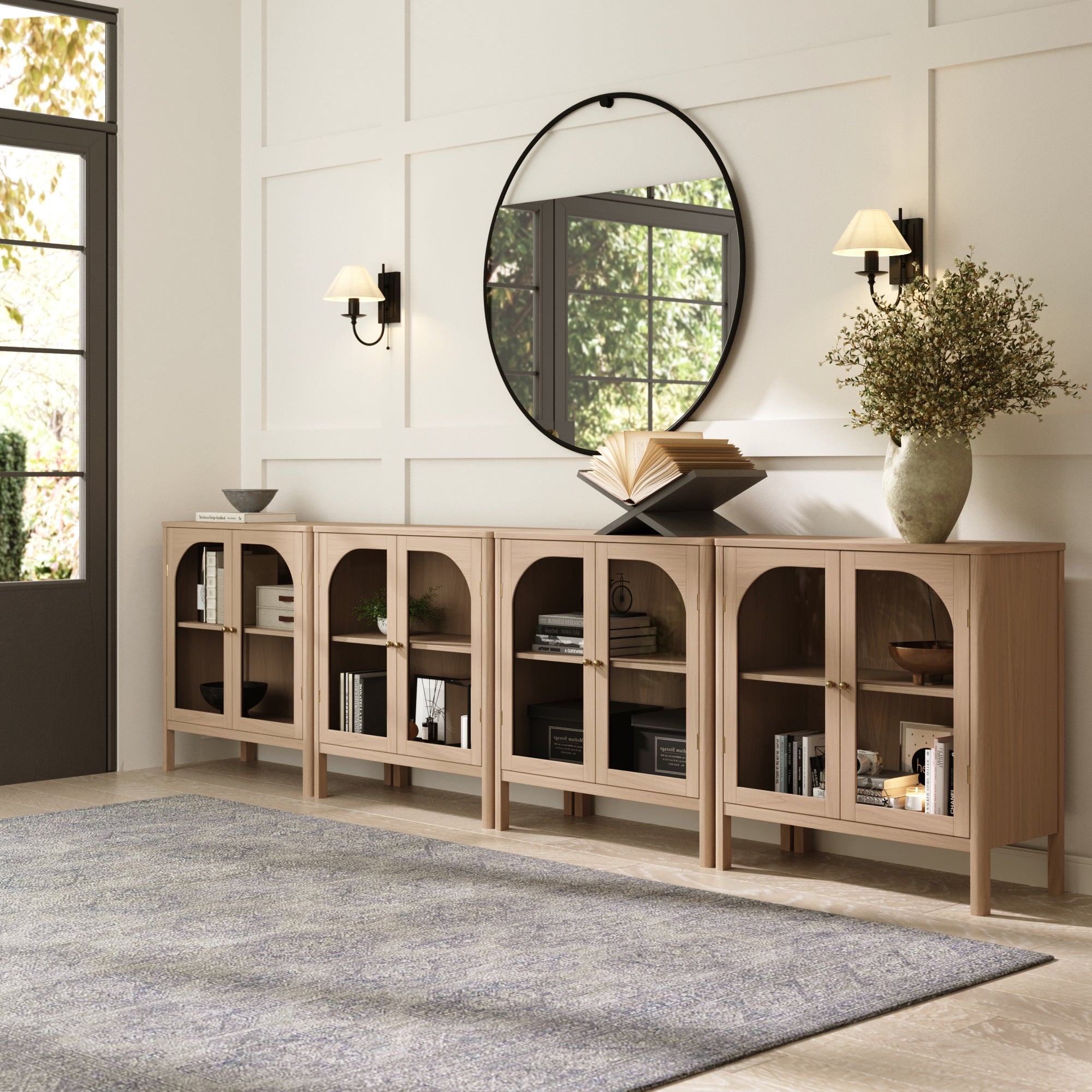 A modern entryway showcases the Nathan James Large Glass Door Curio Cabinet TV Console Credenza in Raw Oak (Set of 4), paired with decorative books, vases, a large round mirror, two wall sconces, and a gray rug on a light floor with sunlight streaming in.