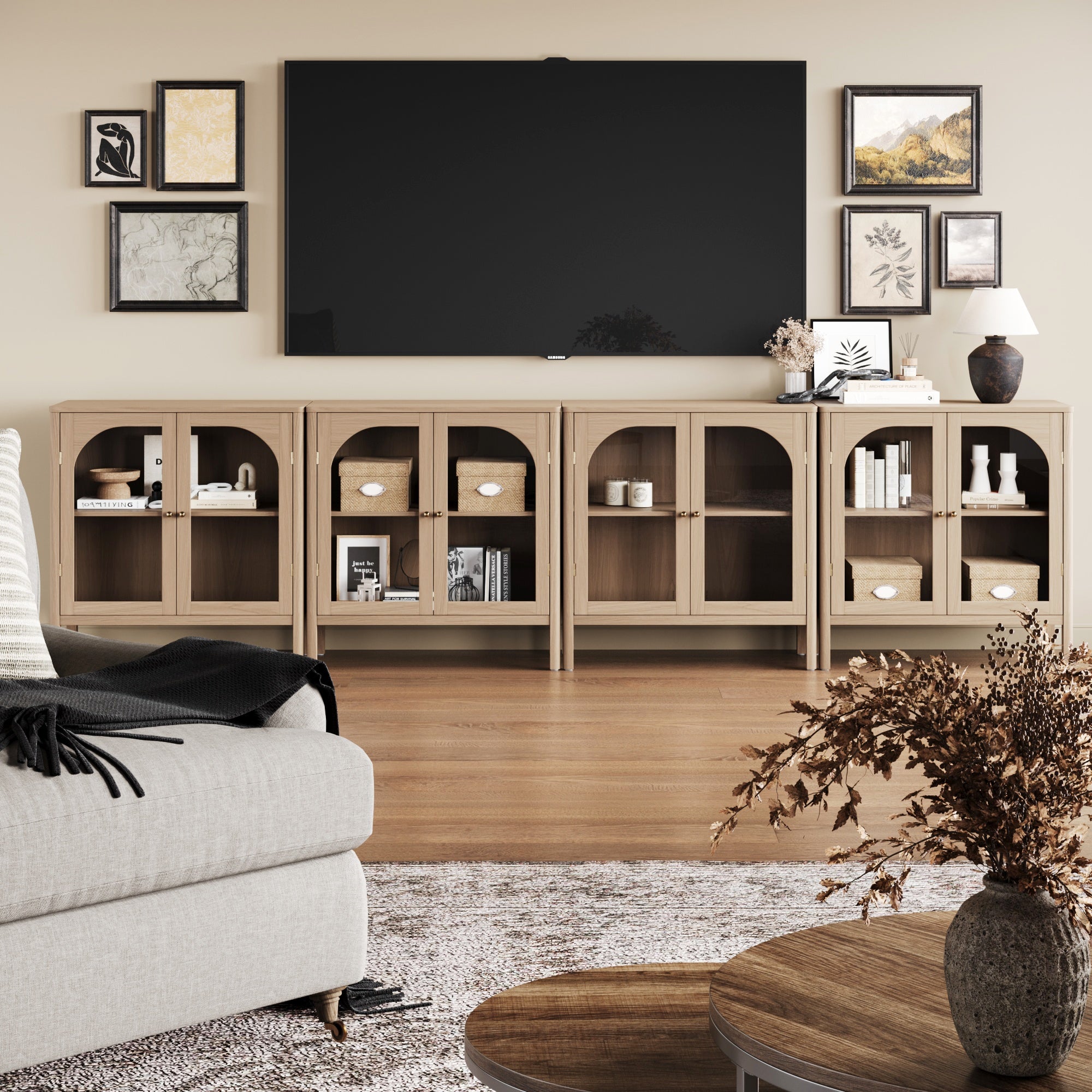 A modern living room featuring the Nathan James Large Glass Door Curio Cabinet TV Console Credenza in Raw Oak (set of 4), a wall-mounted TV, framed art, books, decor, a light sofa, round wood coffee table, and a neutral rug.