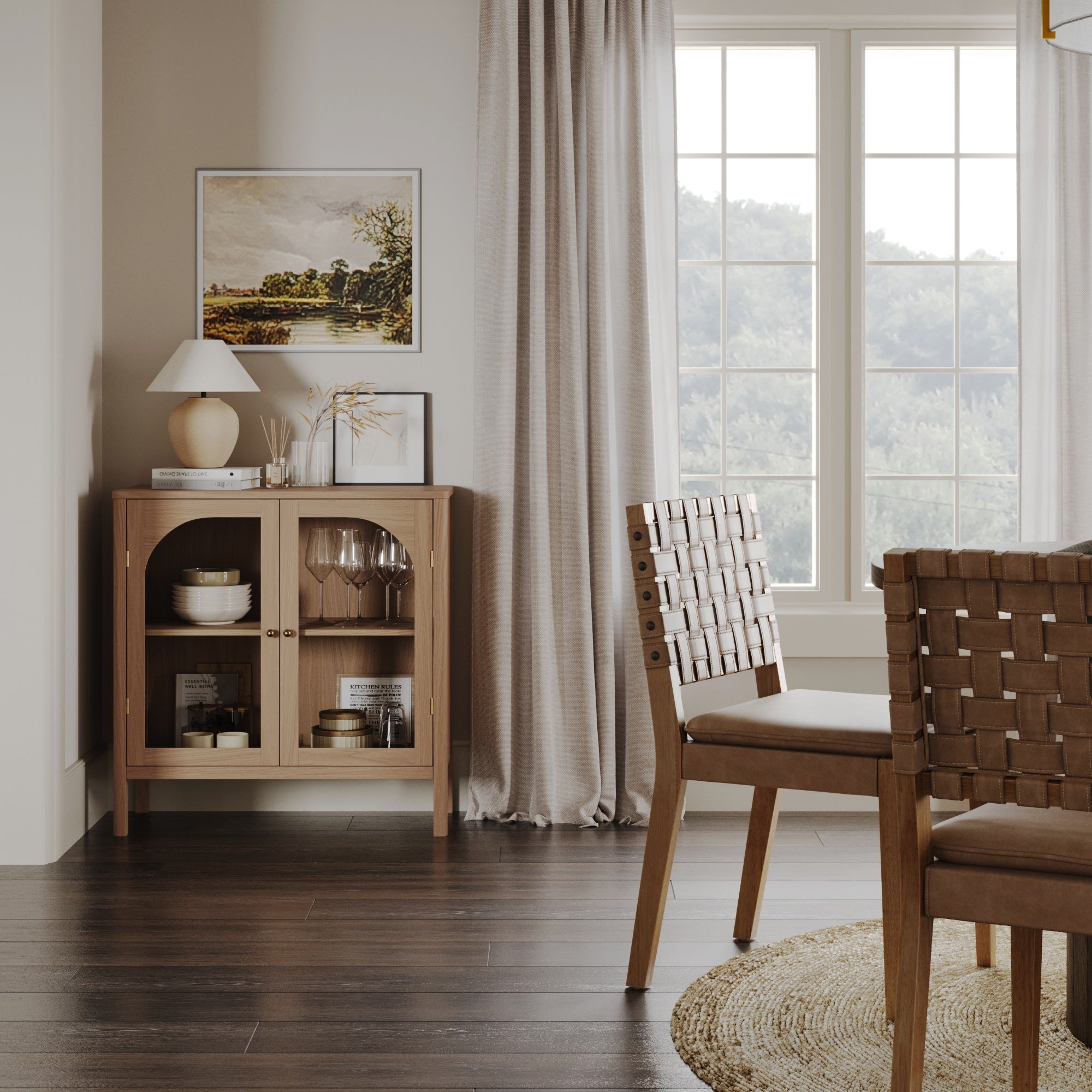A cozy dining room with wooden chairs, a round woven rug, a small cabinet with glass doors, decor items, framed art, and large windows with sheer curtains letting in natural light.