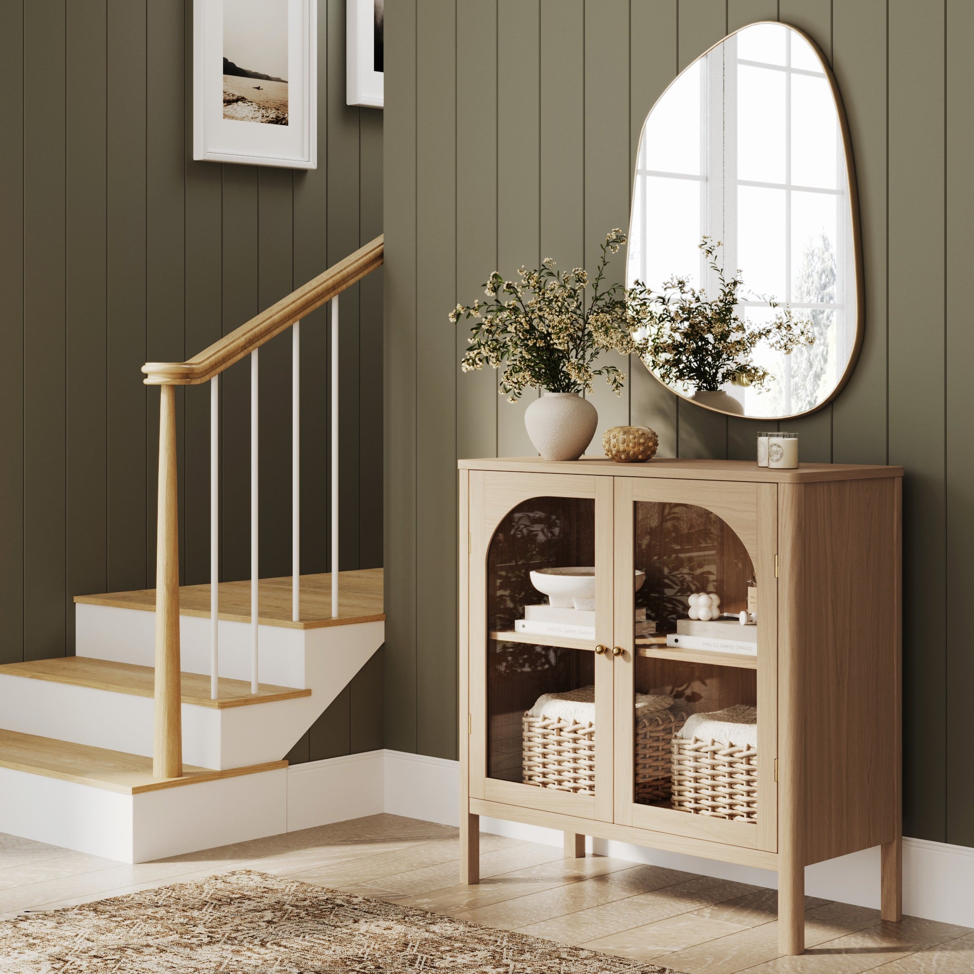 A modern entryway with a light wood cabinet, decorative vases, and baskets. A large asymmetrical mirror hangs above, and wooden stairs with white railings are visible beside paneled olive-green walls.