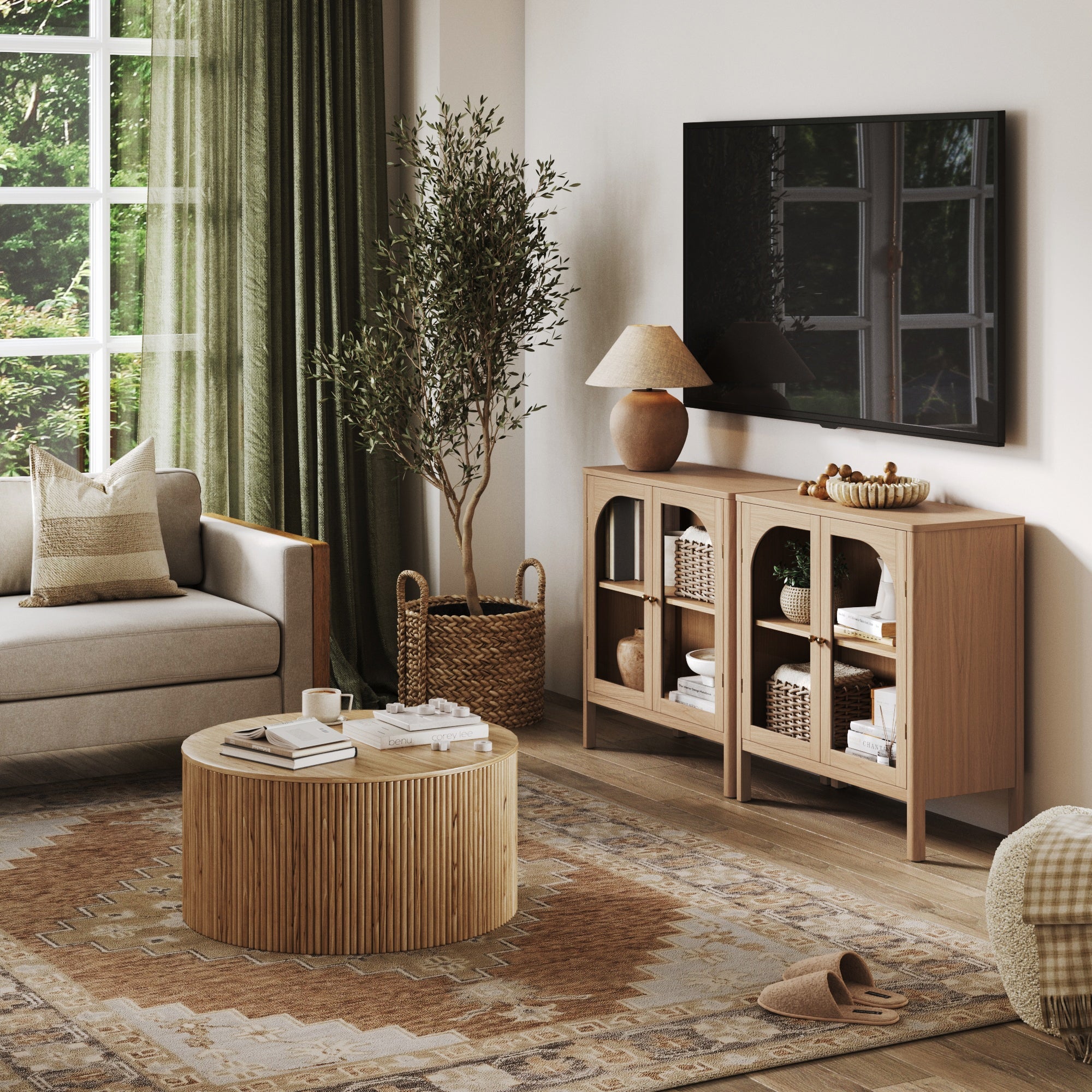 A cozy living room features a round wooden coffee table, beige sofa, patterned rug, and the Nathan James Wood Curio Cabinet with Glass Doors in Raw Oak by the window with green curtains letting in natural light.