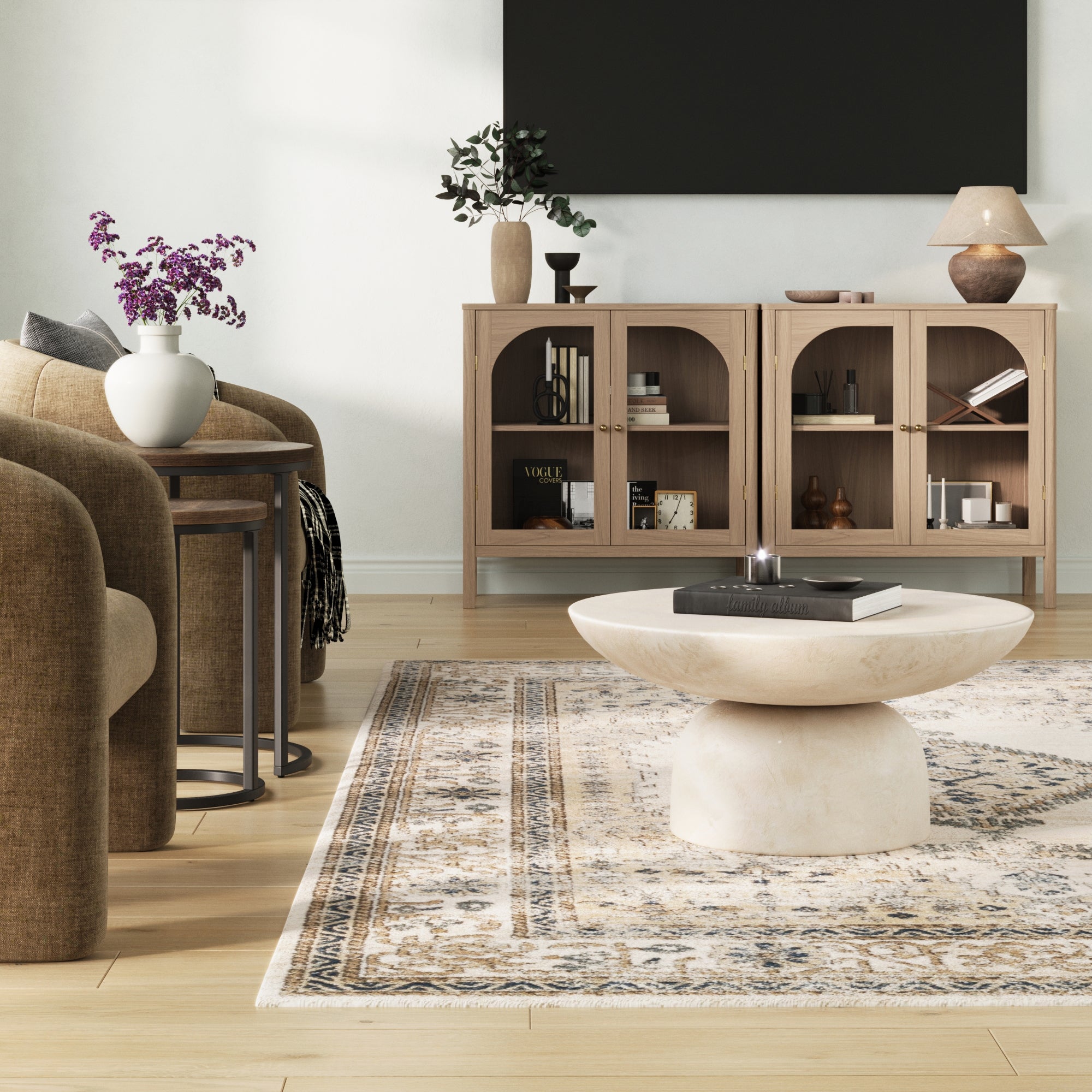 A modern living room features neutral armchairs, a round marble coffee table, patterned rug, and two Nathan James Curio Cabinet TV Stand Credenzas with glass doors in raw oak. Decor includes floral vases, books, and large black wall art.