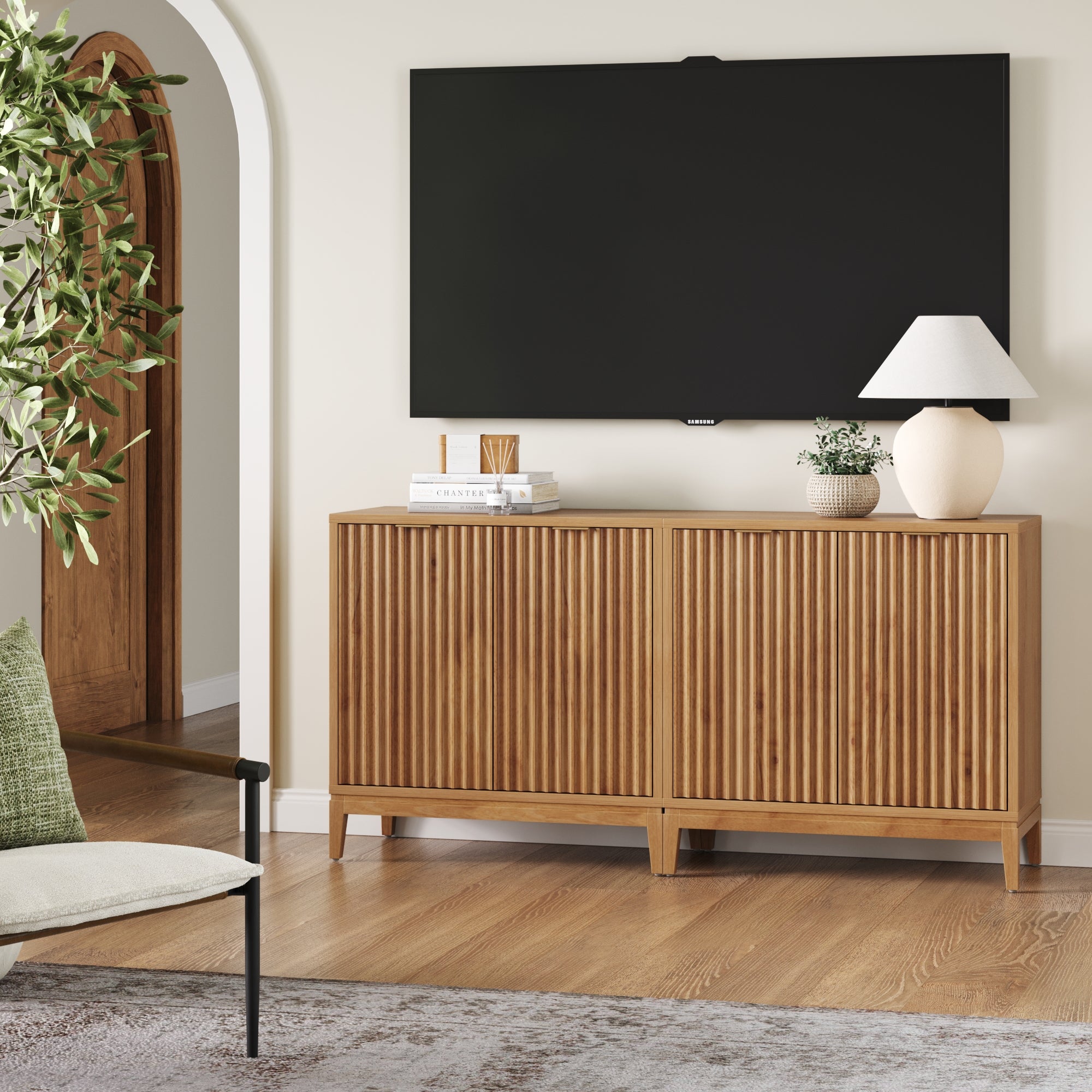 A modern living room showcases the Nathan James Mid-Century Fluted TV Console Credenza in Warm Pine (Set of 2), styled with decorative accents, a white lamp, flat-screen TV, green plant, cushioned chair, and wooden flooring.