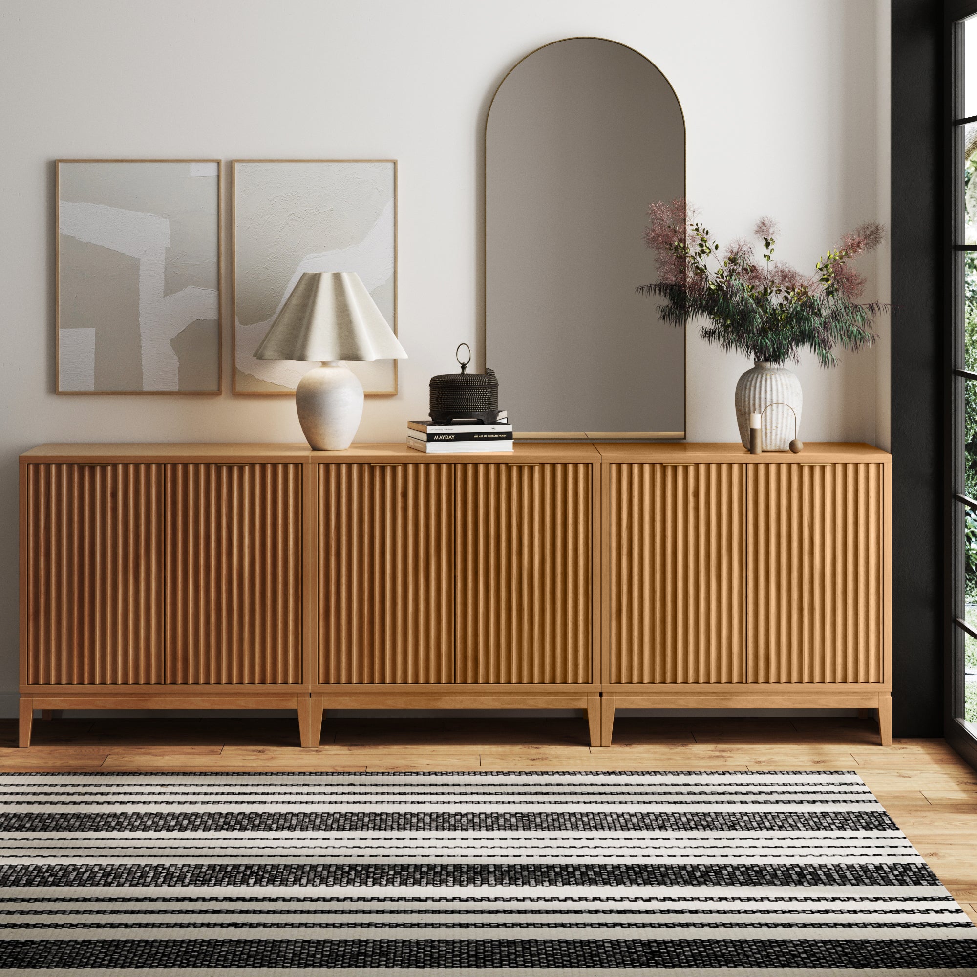 The Nathan James Mid-Century Fluted TV Console Credenza in Warm Pine (Set of 3) features fluted doors and brass handles, styled with a lamp, books, flowers, and decor. Abstract art and an arched mirror hang above, with a striped rug on the hardwood floor.