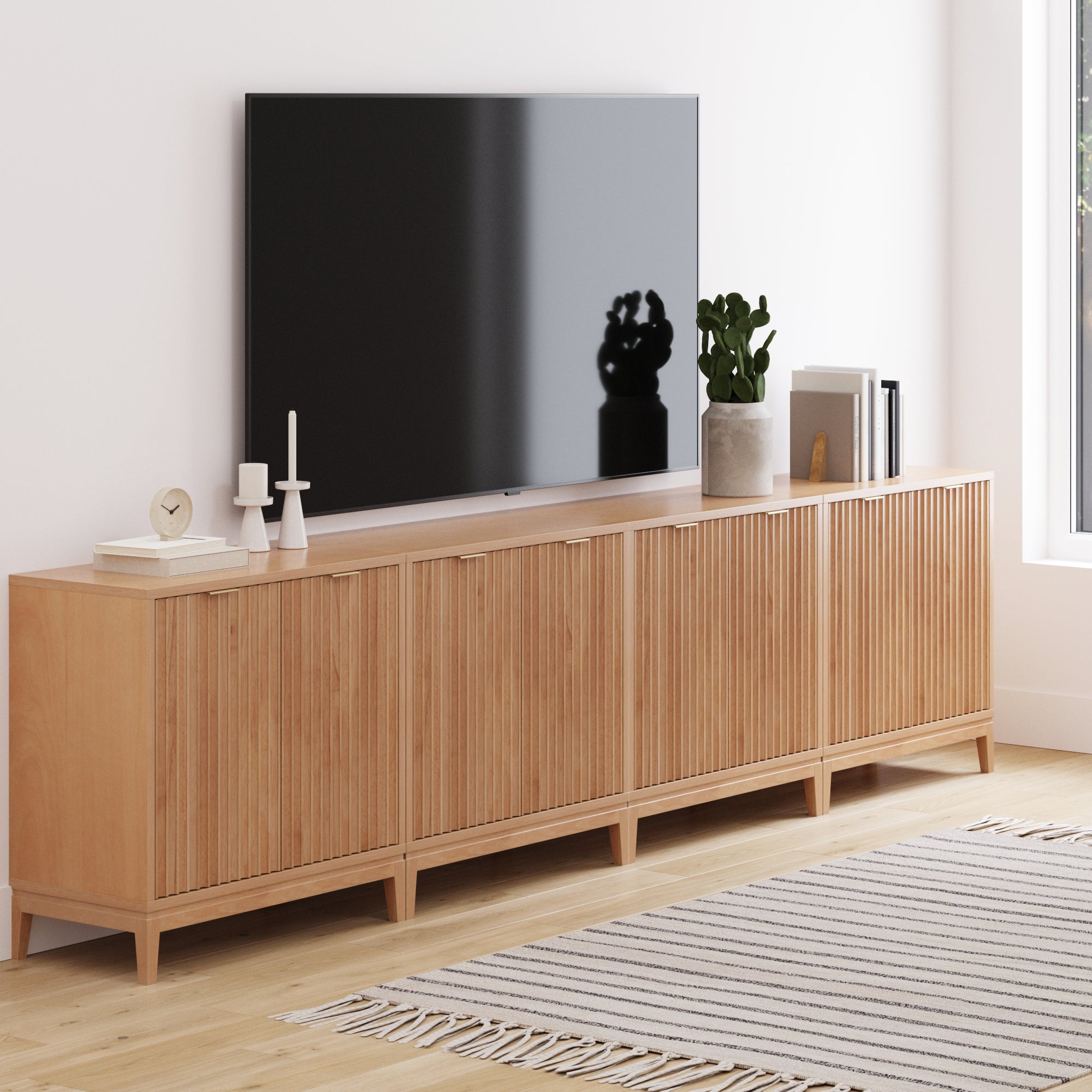 A modern living room features a flat-screen TV on the Nathan James Mid-Century Large Fluted Credenza Console Table in Warm Pine, decorated with candles, clock, potted plant, and books atop a light striped rug on a wooden floor.