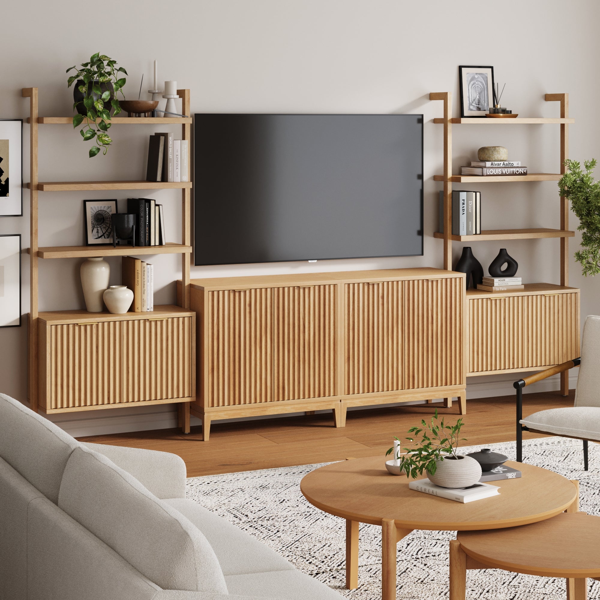 A modern living room with light wood furniture, a Nathan James Mid-Century Entertainment Center in Warm Pine (2 Cabinets), wall-mounted TV, neutral decor, plants, framed art, a round coffee table, and a gray sofa on a patterned rug.