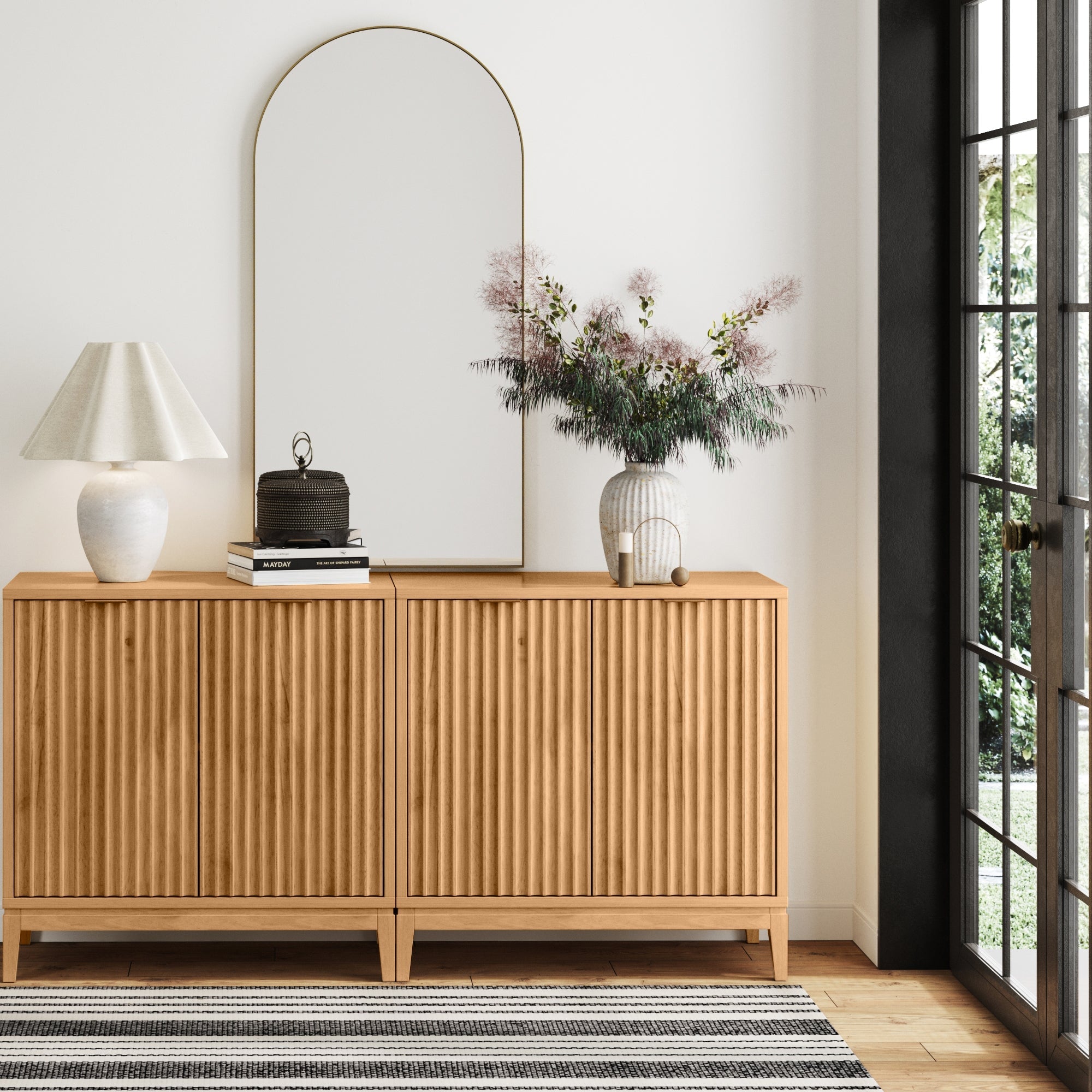 The Nathan James Mid-Century Fluted TV Console Credenza in Warm Pine (Set of 2) stands against a white wall, topped with a lamp, books, decorative box, and vase of flowers. A large arched mirror and window fill the space with natural light.