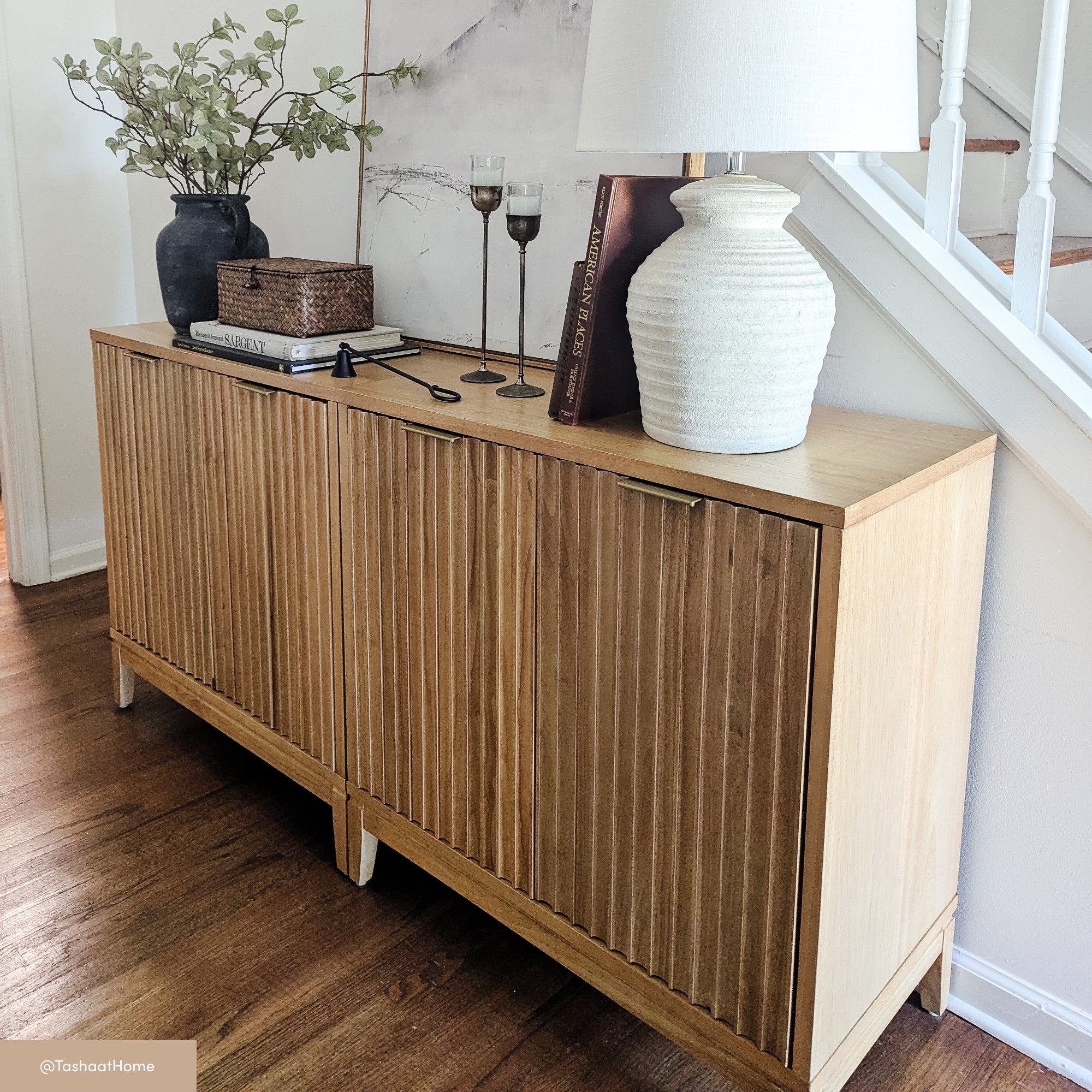 A light wood sideboard with vertical grooves sits against a wall under a staircase. On top are a white lamp, books, a glass vase with greenery, two candle holders, and a pair of glasses.
