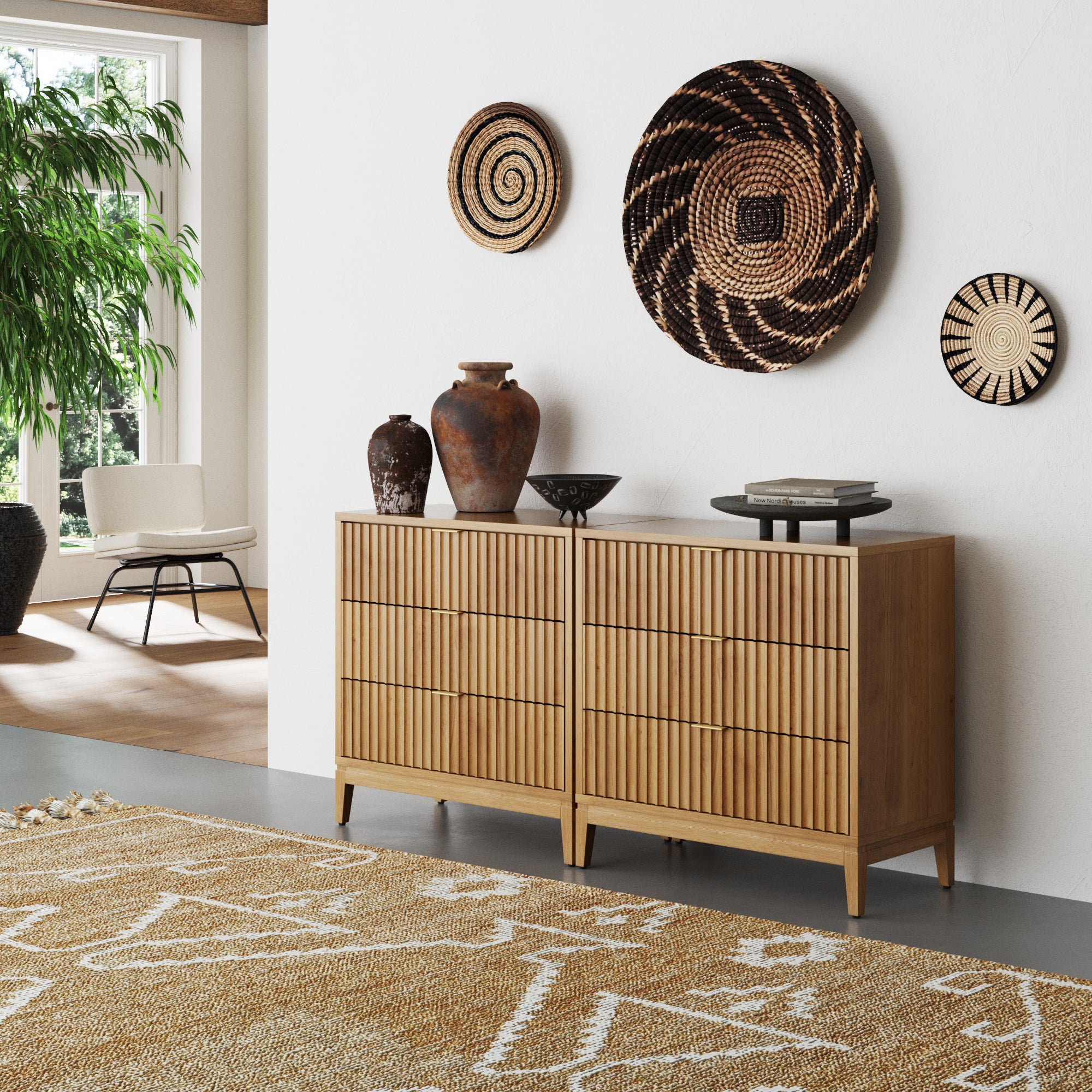 A modern, minimalist living space showcasing the Nathan James Mid-Century Fluted 3-Drawer Dressers in Warm Pine (Set of 2), topped with decorative vases, plus woven wall baskets, a patterned rug, potted plant, and a white chair in the background.