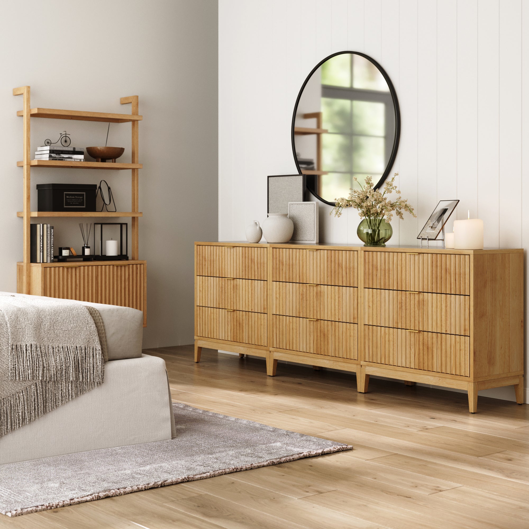 A modern bedroom with light wood floors, a gray rug, a beige sofa, and three Nathan James Mid-Century Fluted 3-Drawer Dressers in Warm Pine topped with decor, a round mirror above, plus a wooden shelf filled with books and decorative items.