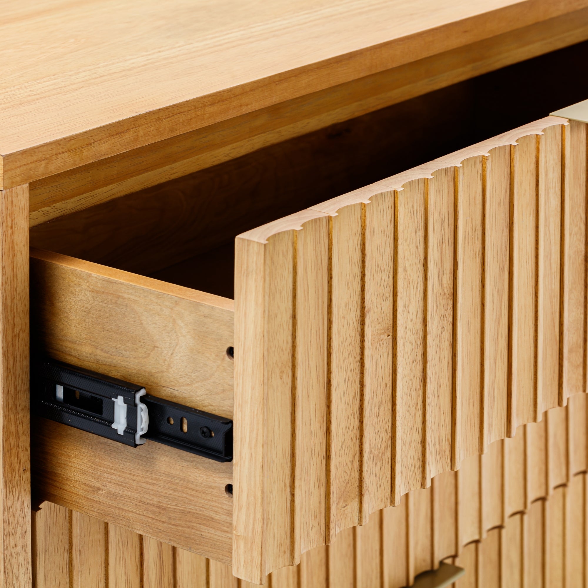 Close-up of a wooden dresser with one drawer partially open, showing a grooved front design and visible black metal sliding mechanism on the side of the drawer.
