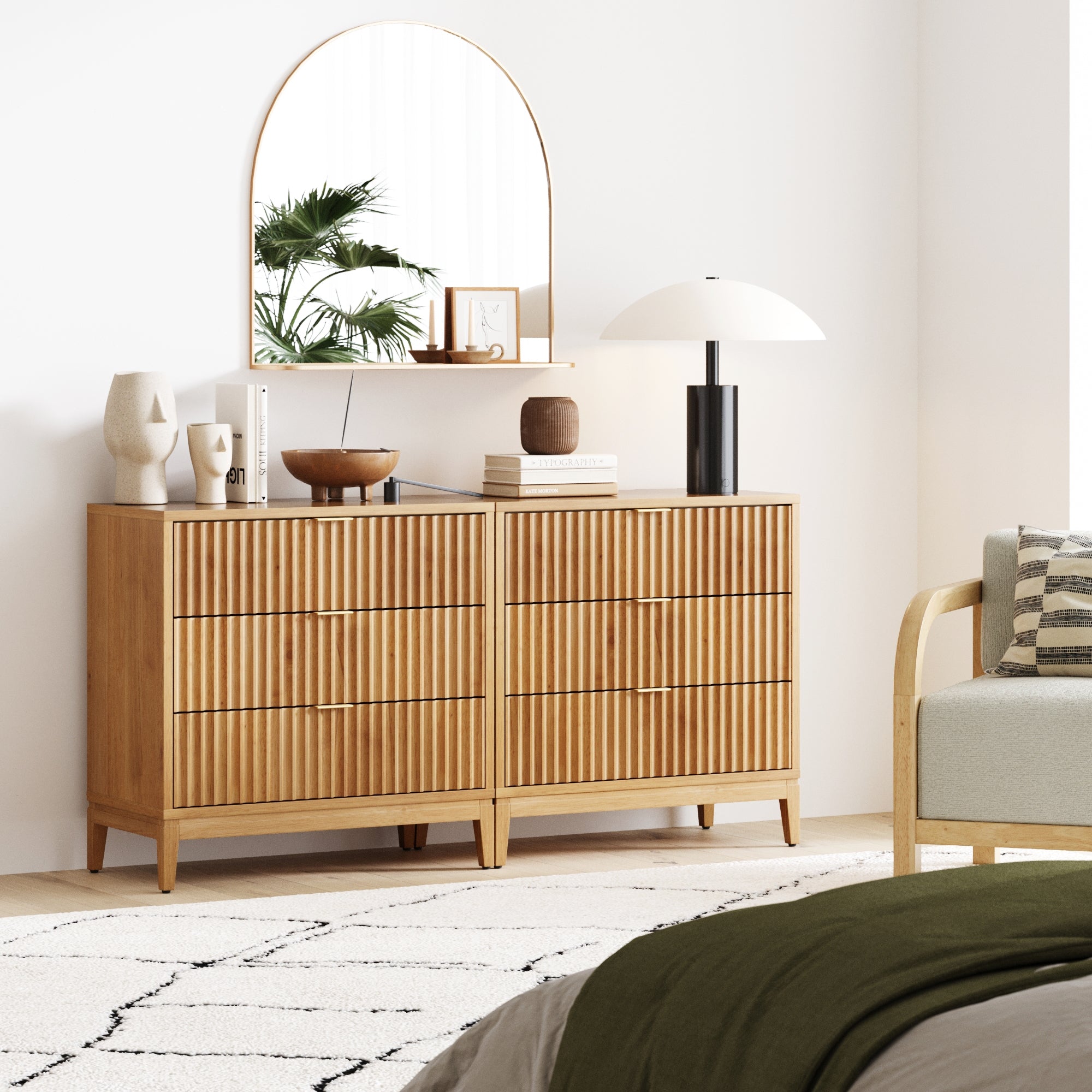 A modern bedroom showcases the Nathan James Mid-Century Fluted 3-Drawer Dresser in Warm Pine with soft-close drawers, styled with decorative pottery, books, a lamp, a mirror reflecting greenery, and a geometric rug for a cozy minimalist vibe.