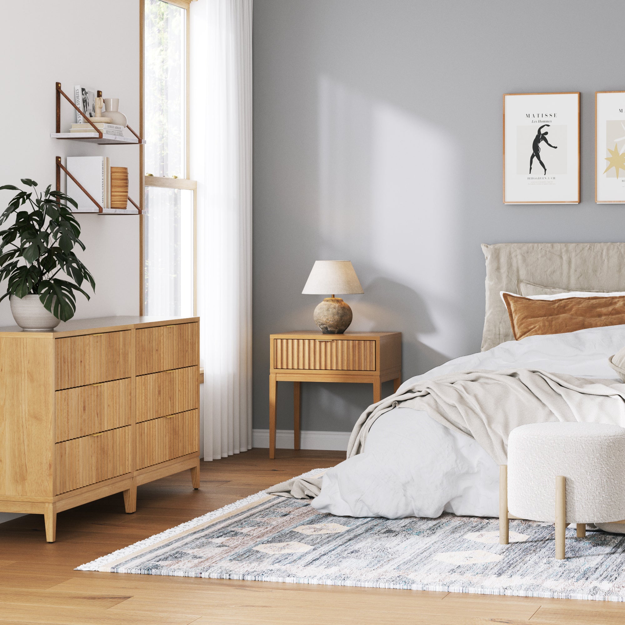 A cozy bedroom with a gray accent wall, wooden furniture, a neatly made bed with neutral bedding, a small nightstand with a lamp, framed art, a potted plant, and sunlight streaming through a window with white curtains.