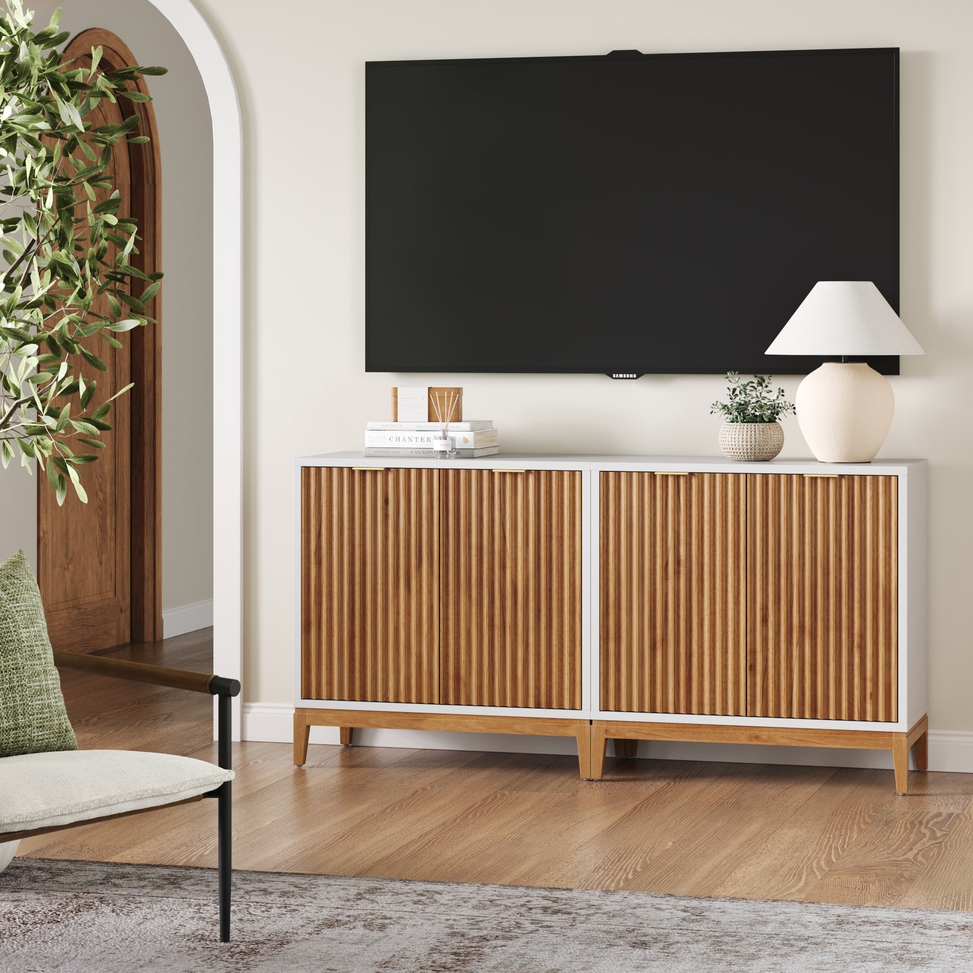 A modern living room highlights the Nathan James Mid-Century Fluted TV Console Credenza in white (set of 2) beneath a wall-mounted TV, styled with decor and a plant. An armchair, neutral walls, and wood flooring complete the space’s inviting look.